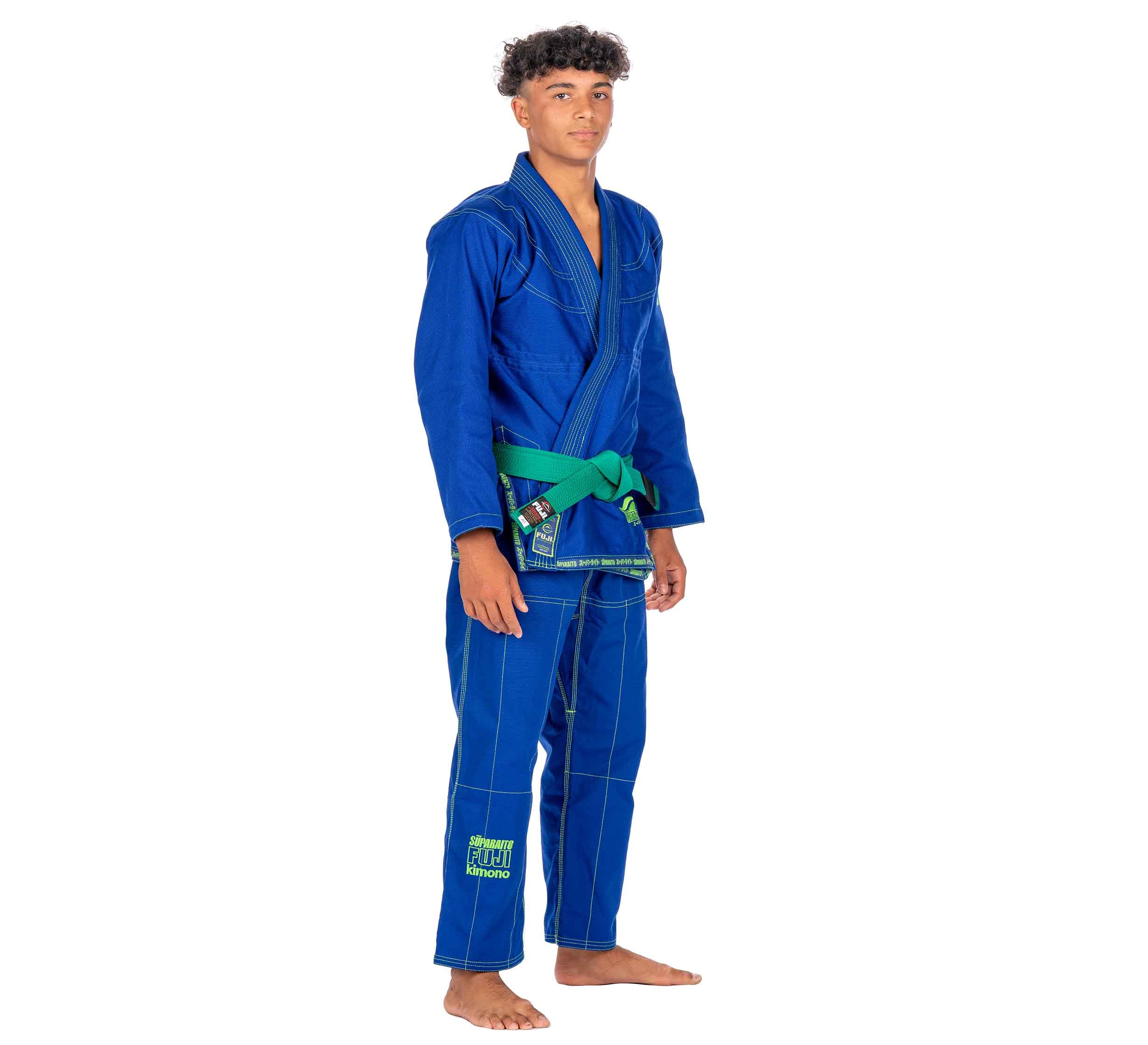 A young man stands barefoot on a white background, facing forward with hands relaxed at his sides, wearing the Suparaito Kids Blue BJJ Gi and a green belt.