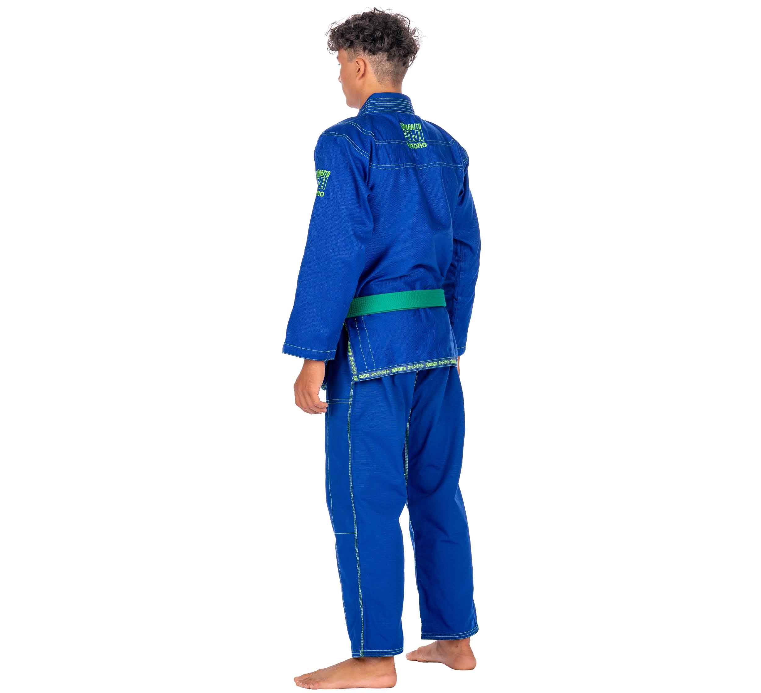 A child with short curly hair stands barefoot, facing away, wearing the Suparaito Kids Blue BJJ Gi featuring green detailing, a green belt, and patches with embroidery on the back and upper arm.