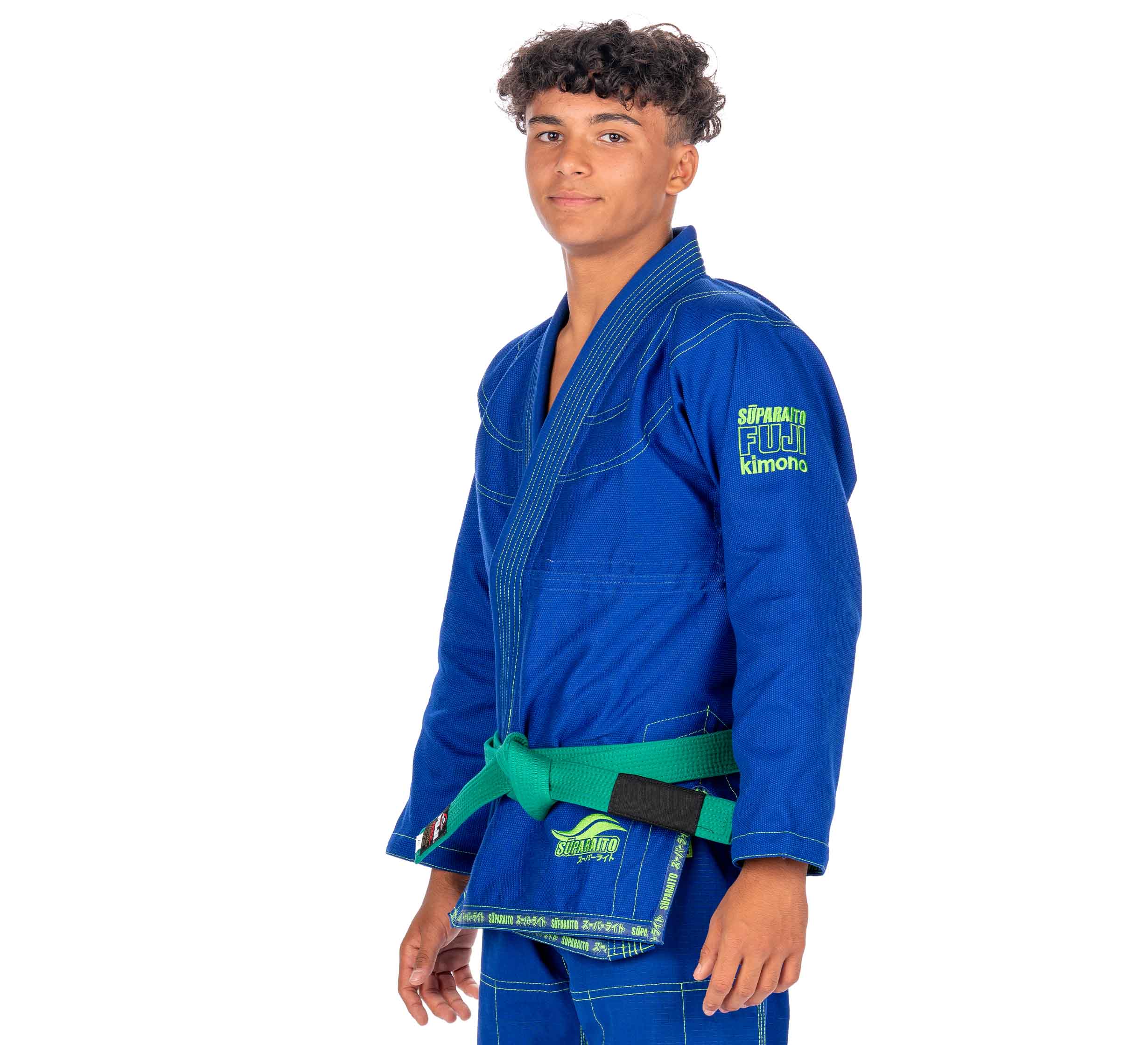 A young person with short curly hair wears the Suparaito Kids Blue BJJ Gi and a green belt, standing slightly angled on a white background while looking at the camera.