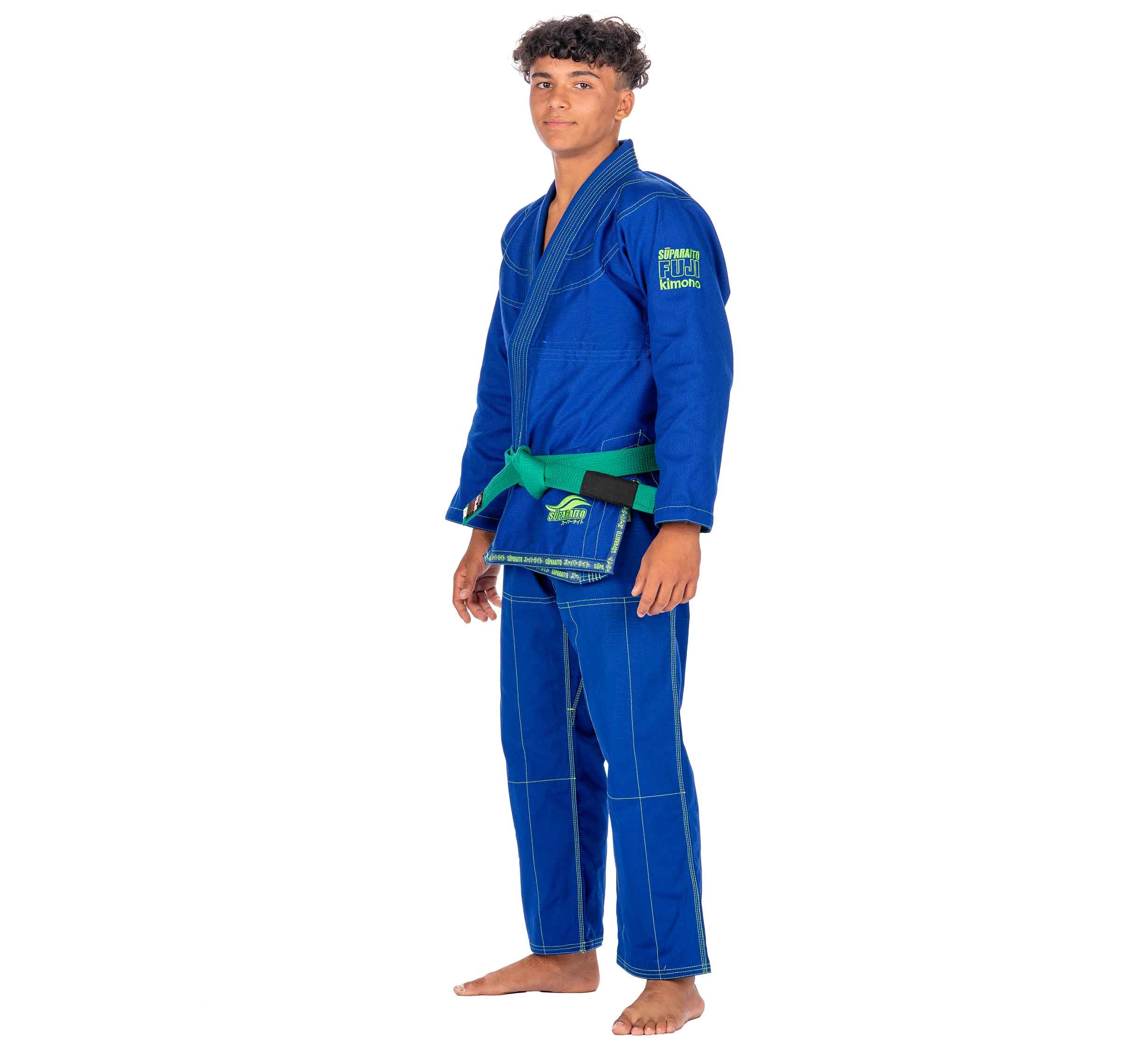 A young boy stands barefoot on a white background, wearing the Suparaito Kids Blue BJJ Gi with a green belt and looking at the camera with a neutral expression.