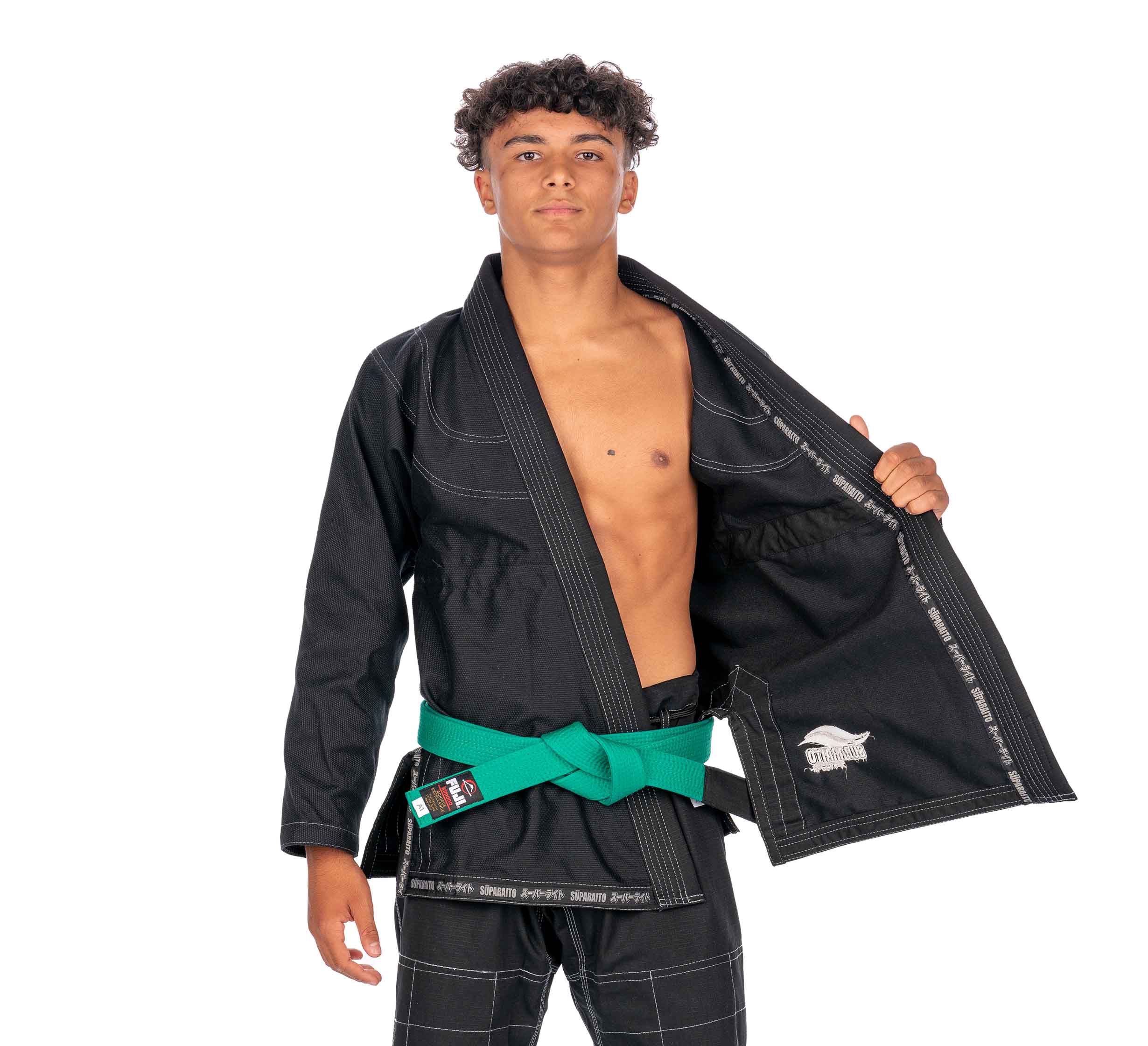 A confident young man with short curly hair wears the Suparaito Kids Black BJJ Gi and a green belt. He stands facing forward, holding open one side of his gi jacket to show the inside.