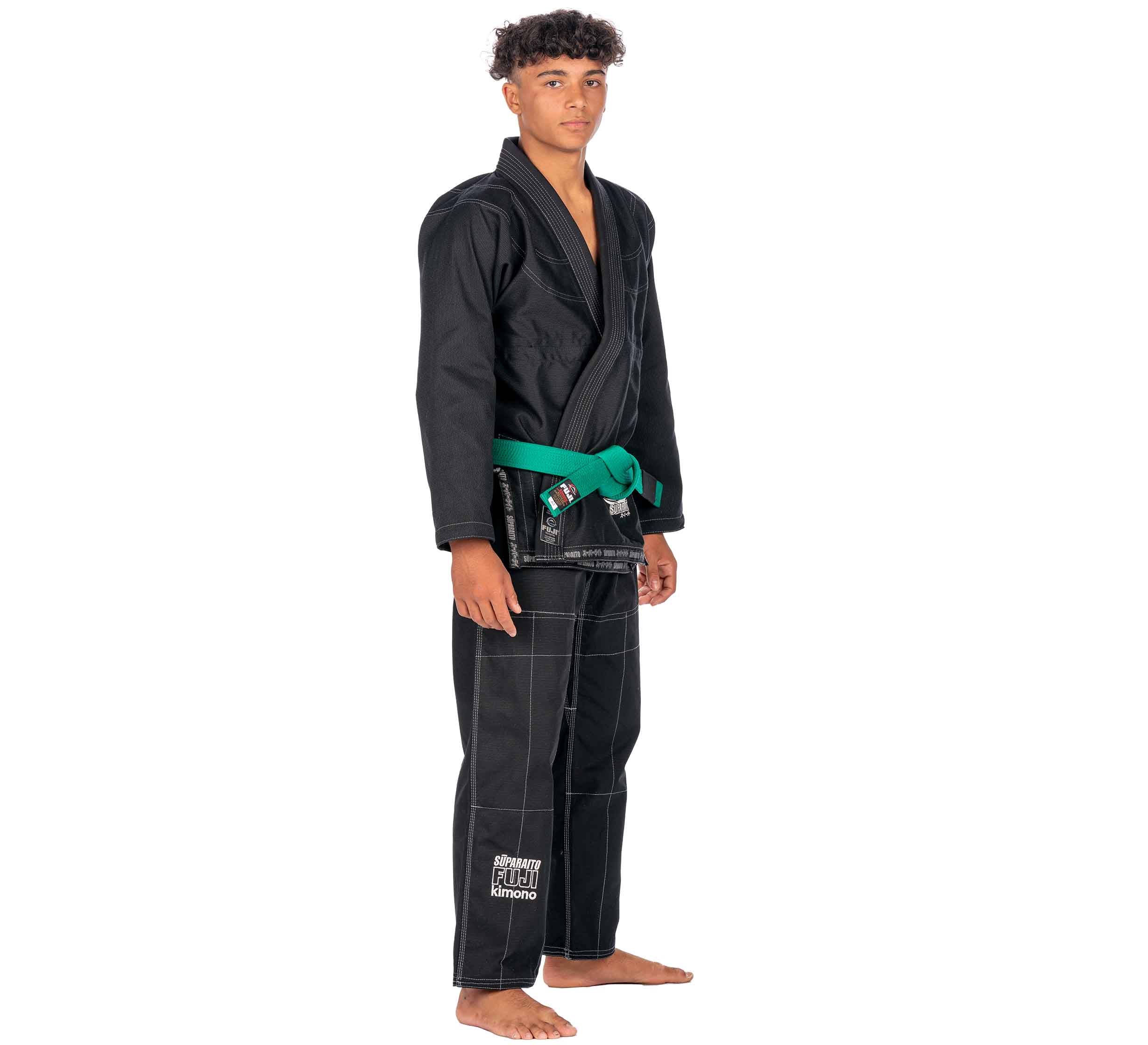 A young person stands barefoot in the Suparaito Kids Black BJJ Gi with a green belt, white stitching, and embroidered text on one leg. They face forward with a neutral expression.