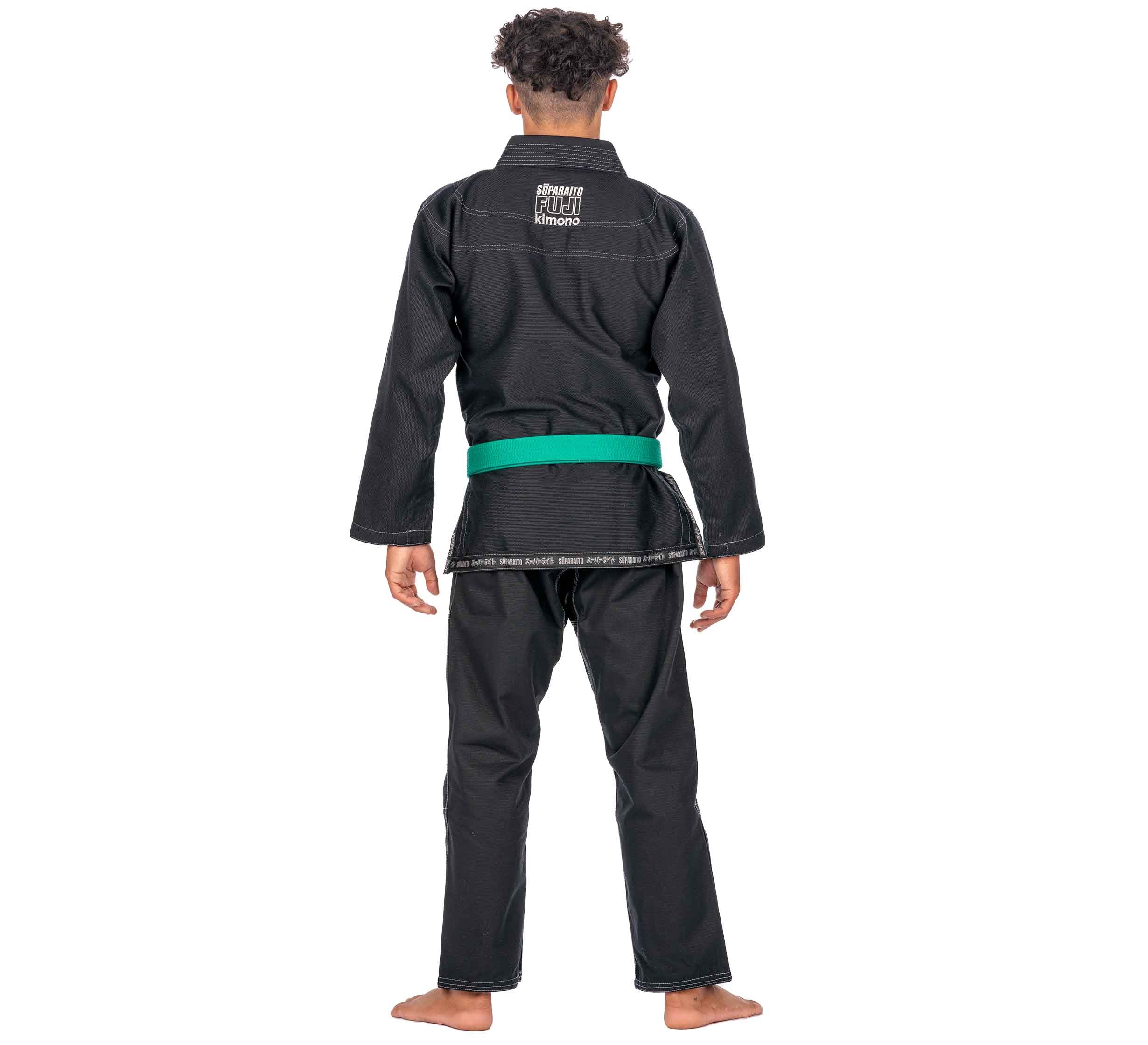 A child stands barefoot, facing away from the camera in a Suparaito Kids Black BJJ Gi with a green belt, set against a white background.