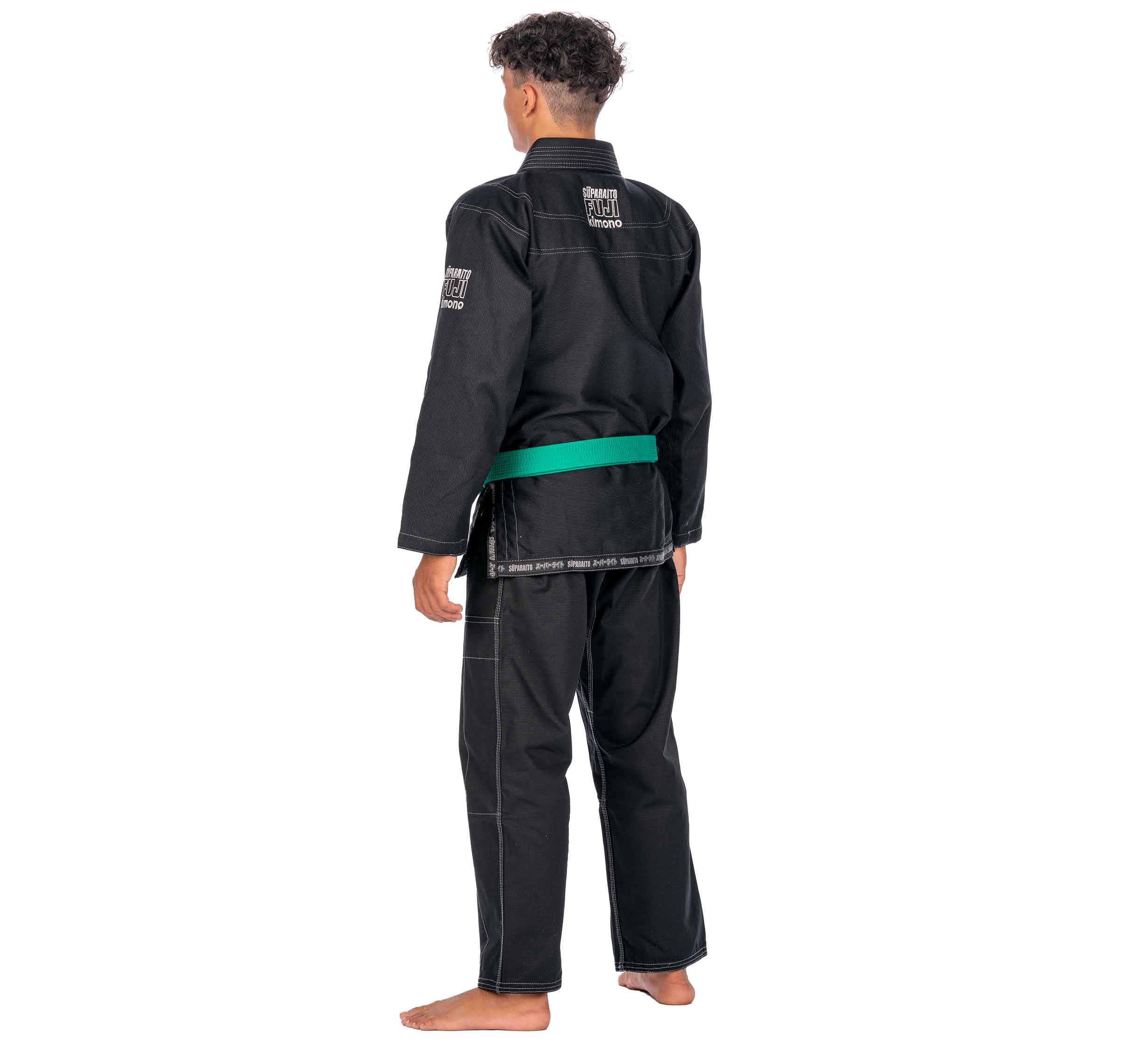 A child stands barefoot facing away, wearing the Suparaito Kids Black BJJ Gi with a green belt. The gi features white stitching and sleeve patches. The background is plain white.