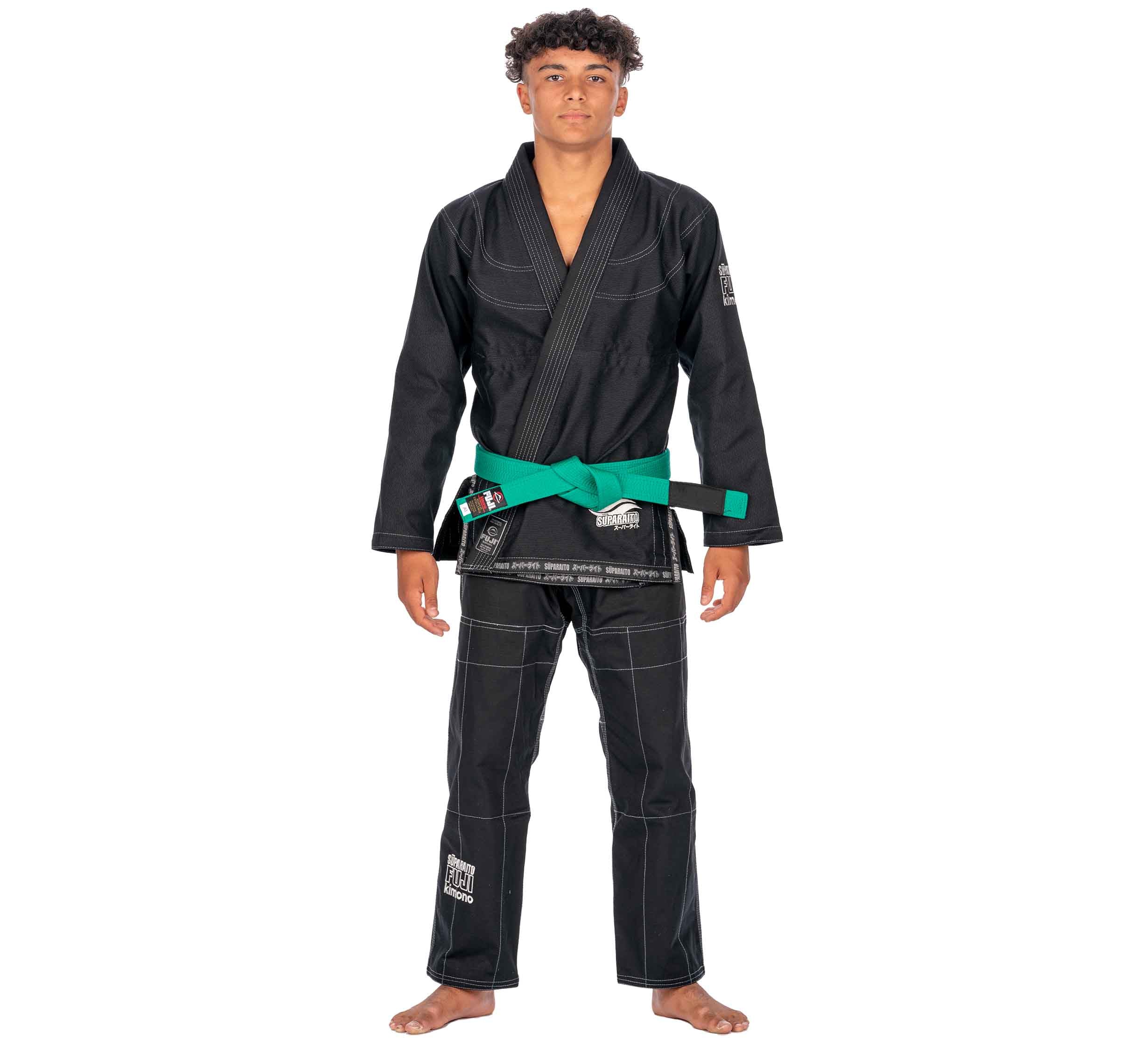 A young man stands barefoot, facing forward in a Suparaito Kids Black BJJ Gi with white stitching and a green belt, set against a plain white background.