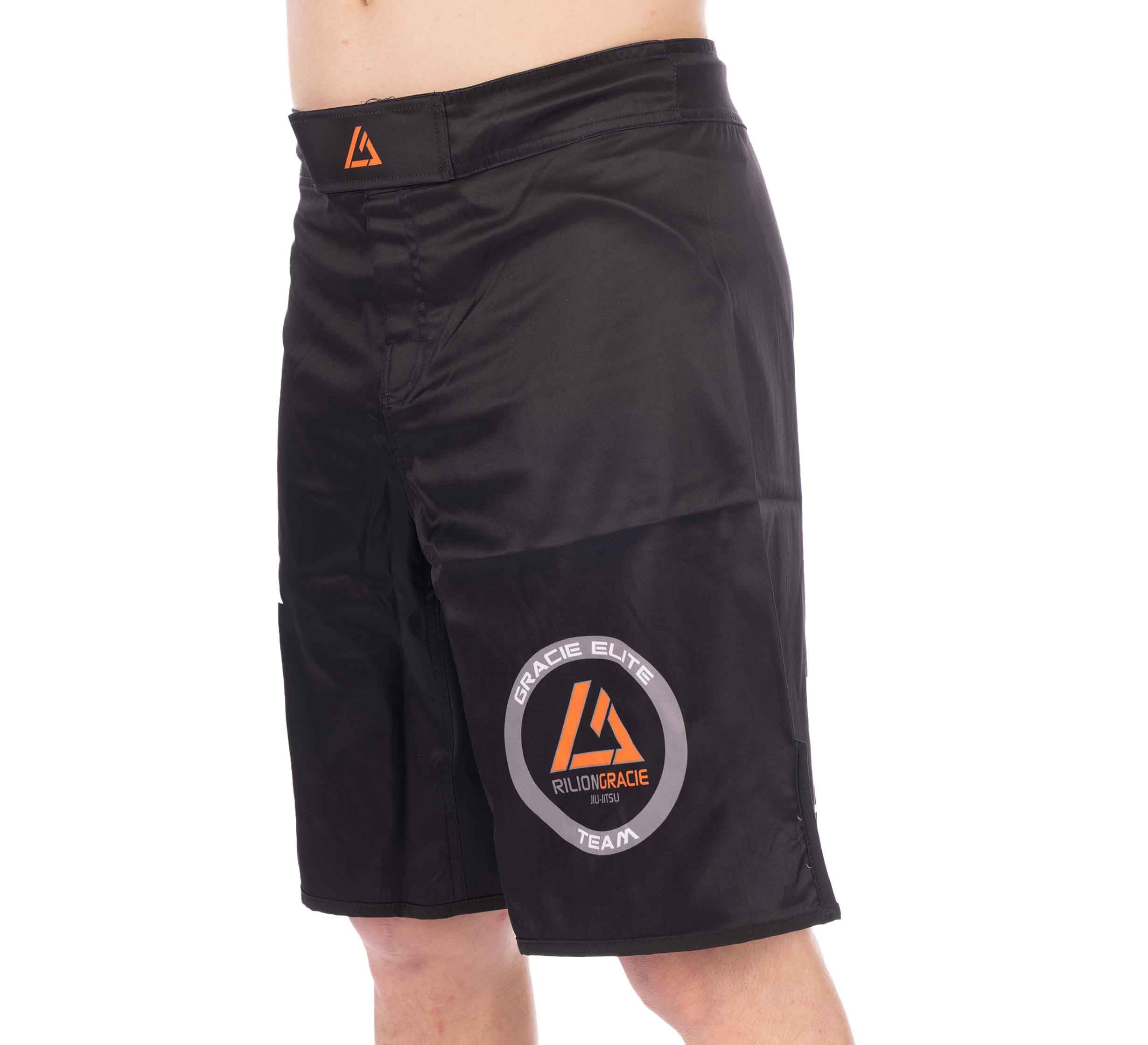Person wearing Rilion Gracie Black Fight Shorts featuring an orange triangle logo on the waistband and a round Gracie Elite Team logo on the left leg. Photo is cropped from waist to just above the knee.