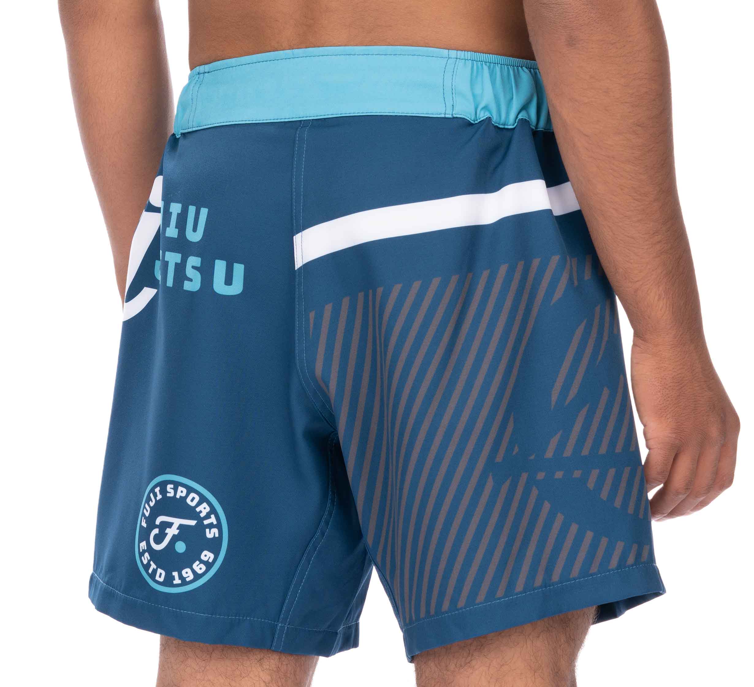 A man wearing Script Grappling Shorts Blue, featuring a light blue waistband, white stripes, and "FUJI SPORTS ESTD 1969" printed on the side.