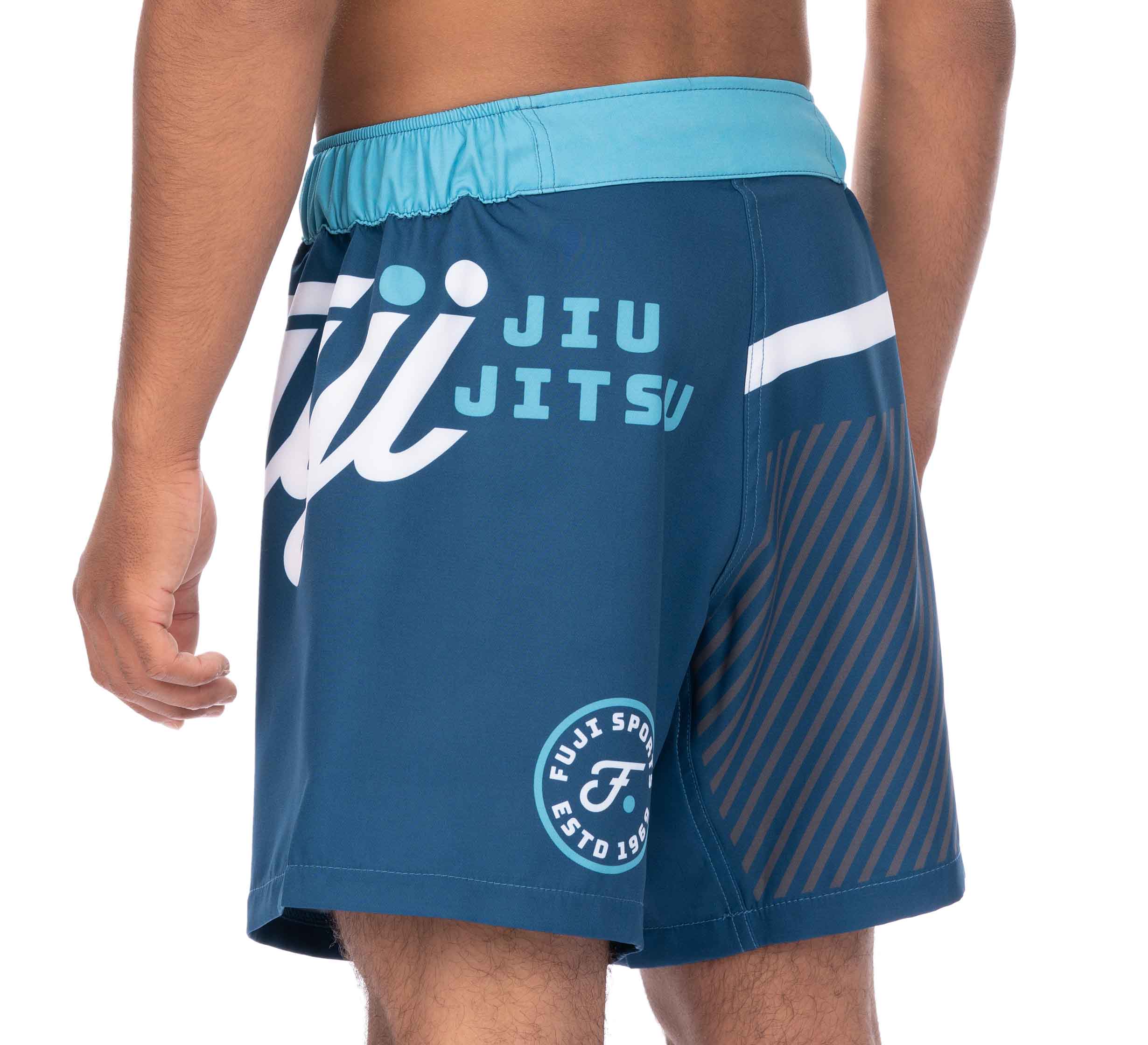A person is seen from behind at an angle wearing Script Grappling Shorts Blue, featuring Jiu Jitsu and Fuji Sports logos on the side, a light blue waistband, and striped detailing.