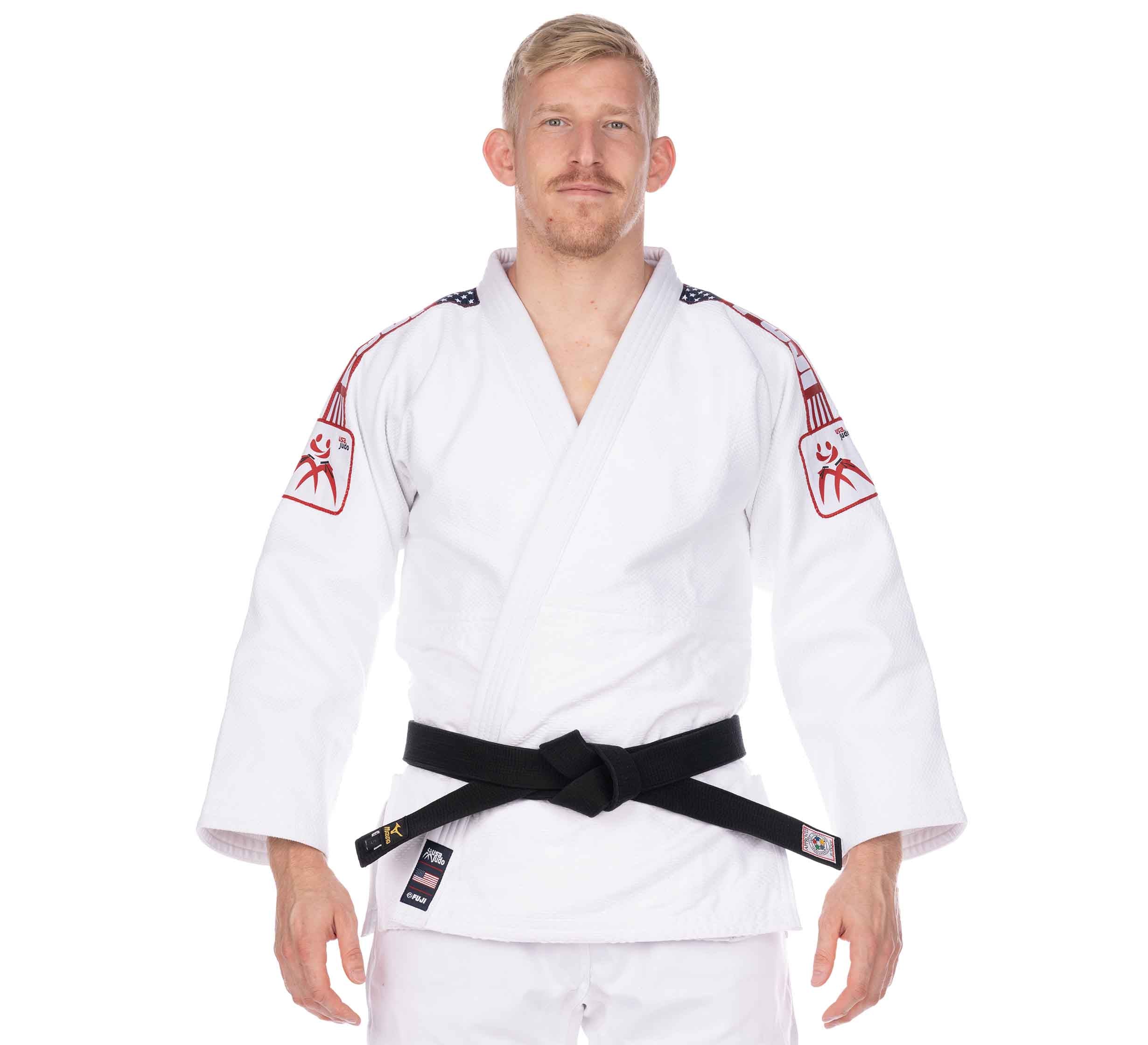 A man with short blond hair and a trimmed beard stands facing forward, wearing the USA Judo Single Weave Gi 2.0 White with red and white shoulder patches, a black belt, and a neutral expression.