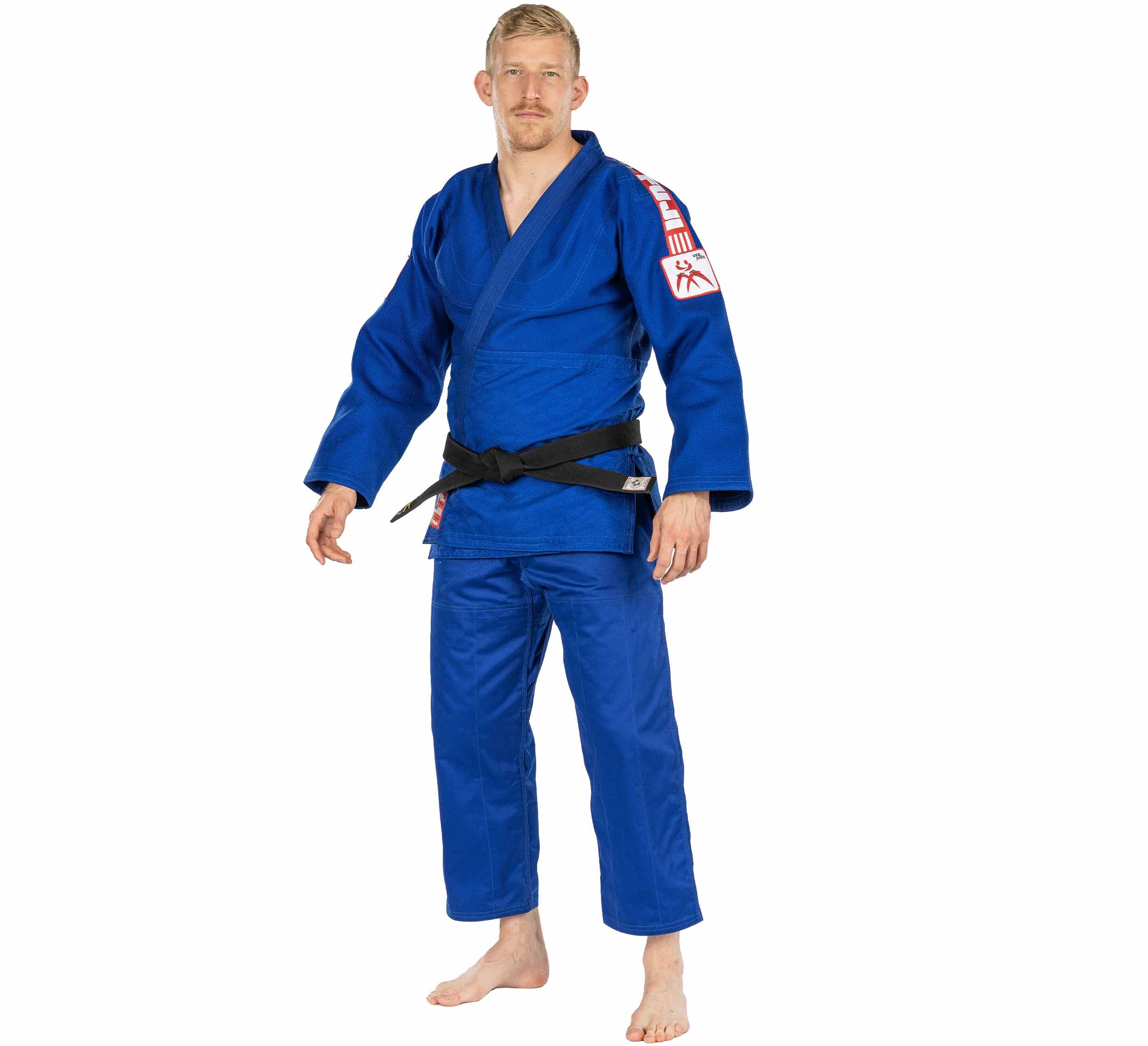 A man stands barefoot in a blue USA Judo Double Weave Gi 2.0 with a black belt, featuring red and white shoulder patches, against a white background.