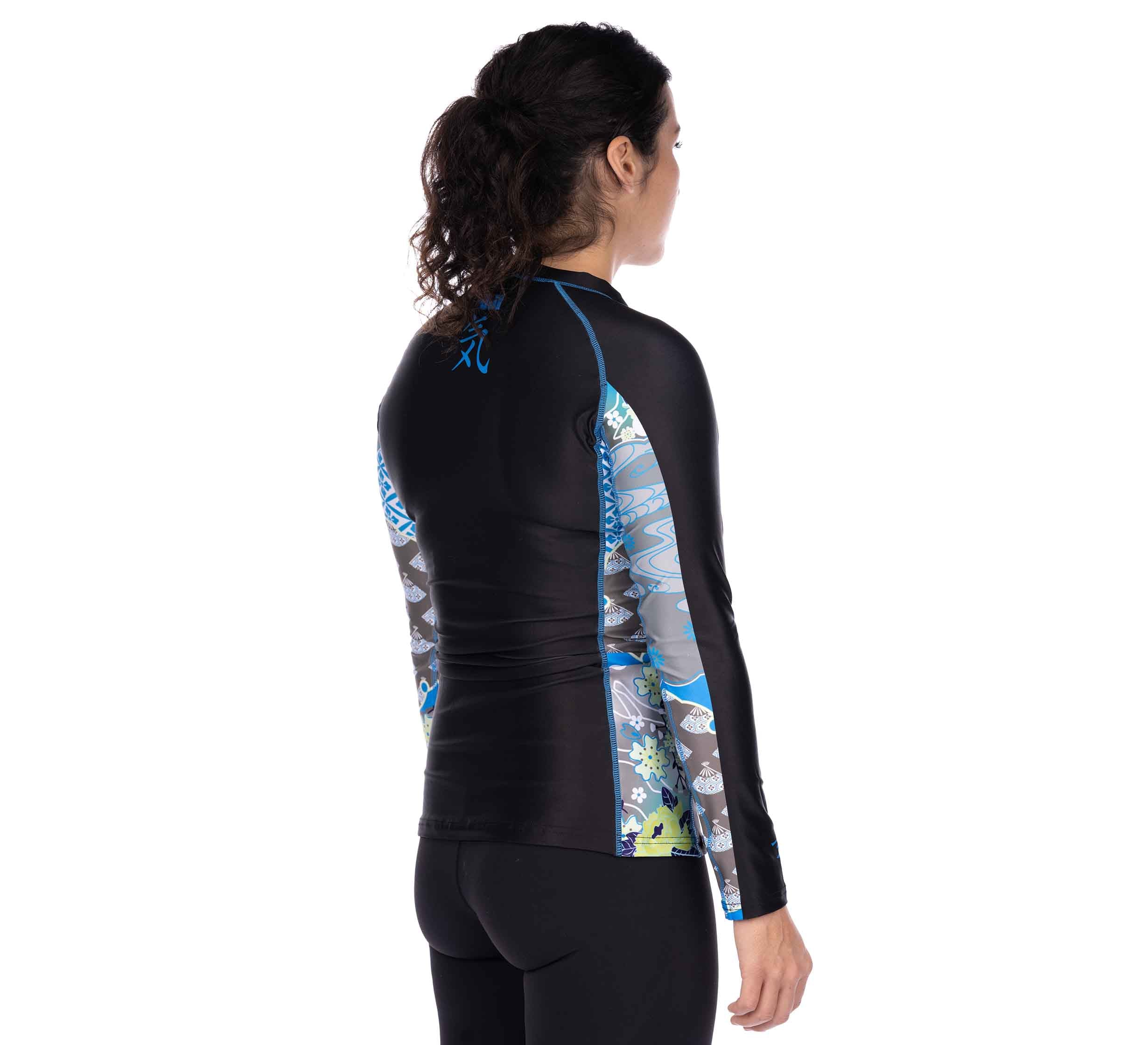 A woman with dark, curly hair in a ponytail, seen from the back, wears the Kimono Womens Long Sleeve Rashguard Blue—an athletic top with fitted black and blue panels and patterned sides.