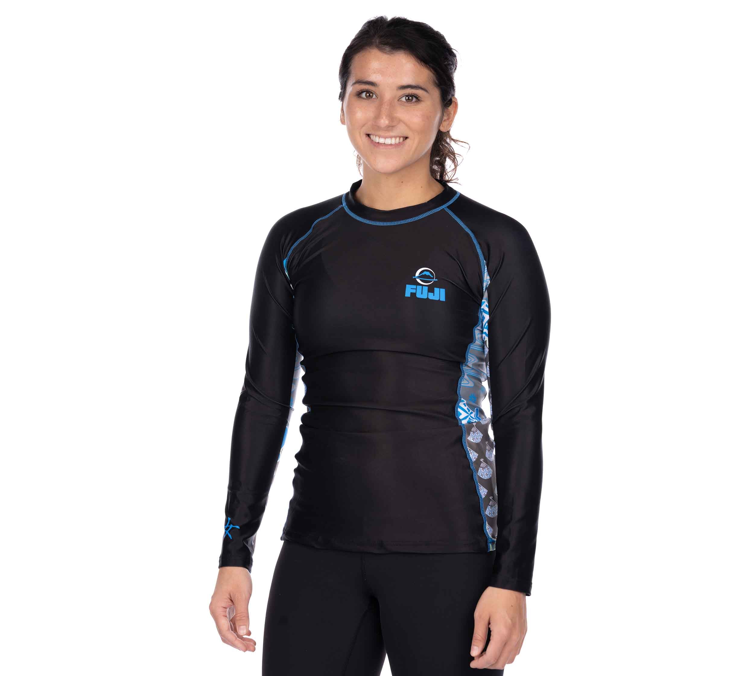 A woman with dark hair tied back is smiling against a white background, wearing the Kimono Womens Long Sleeve Rashguard Blue by FUJI, a black athletic top with blue accents and the FUJI logo on the chest.