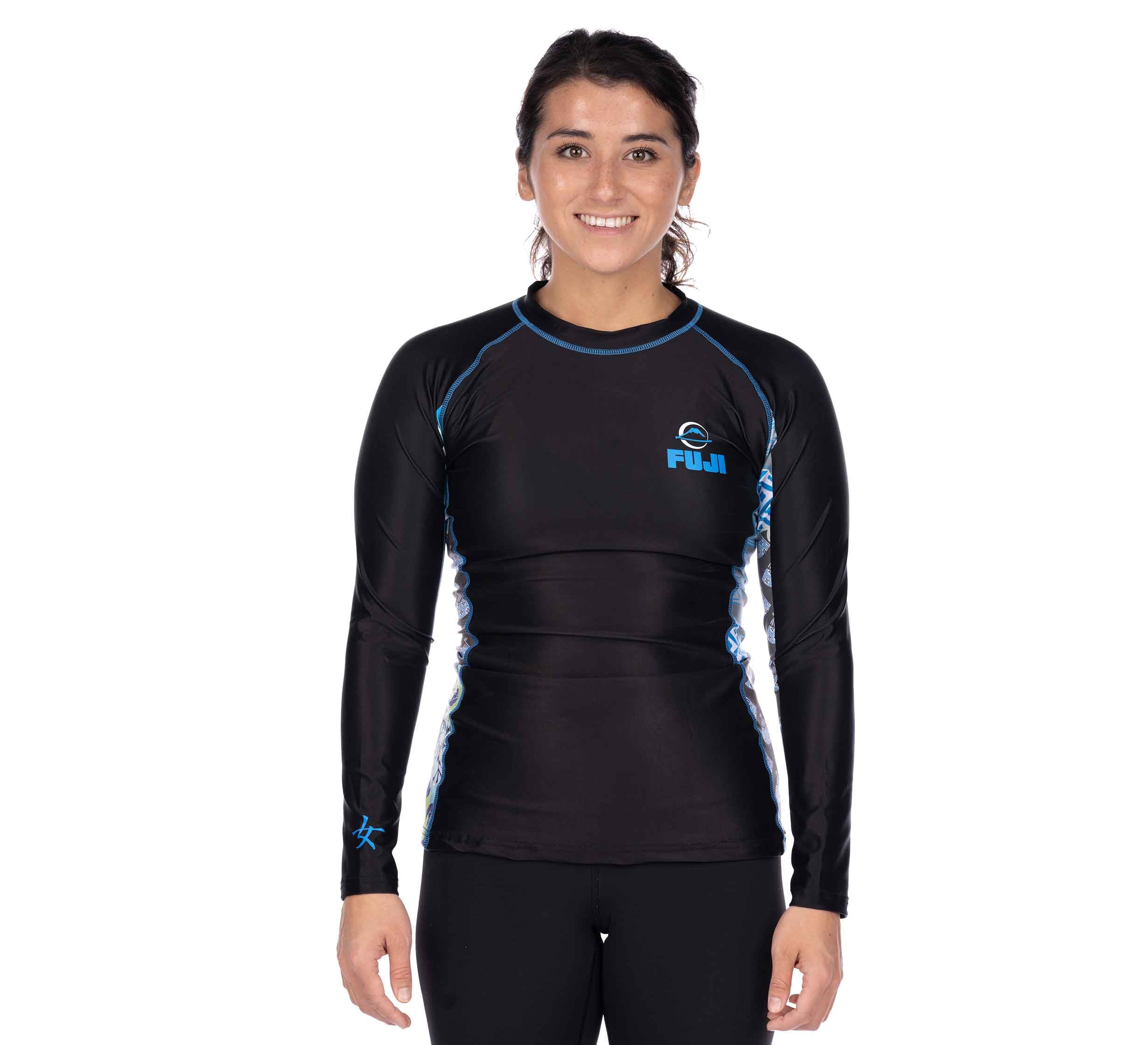 A woman with dark hair pulled back smiles while wearing the "Kimono Womens Long Sleeve Rashguard Blue"—a black rash guard with blue trim and FUJI printed on the chest—against a plain white background.