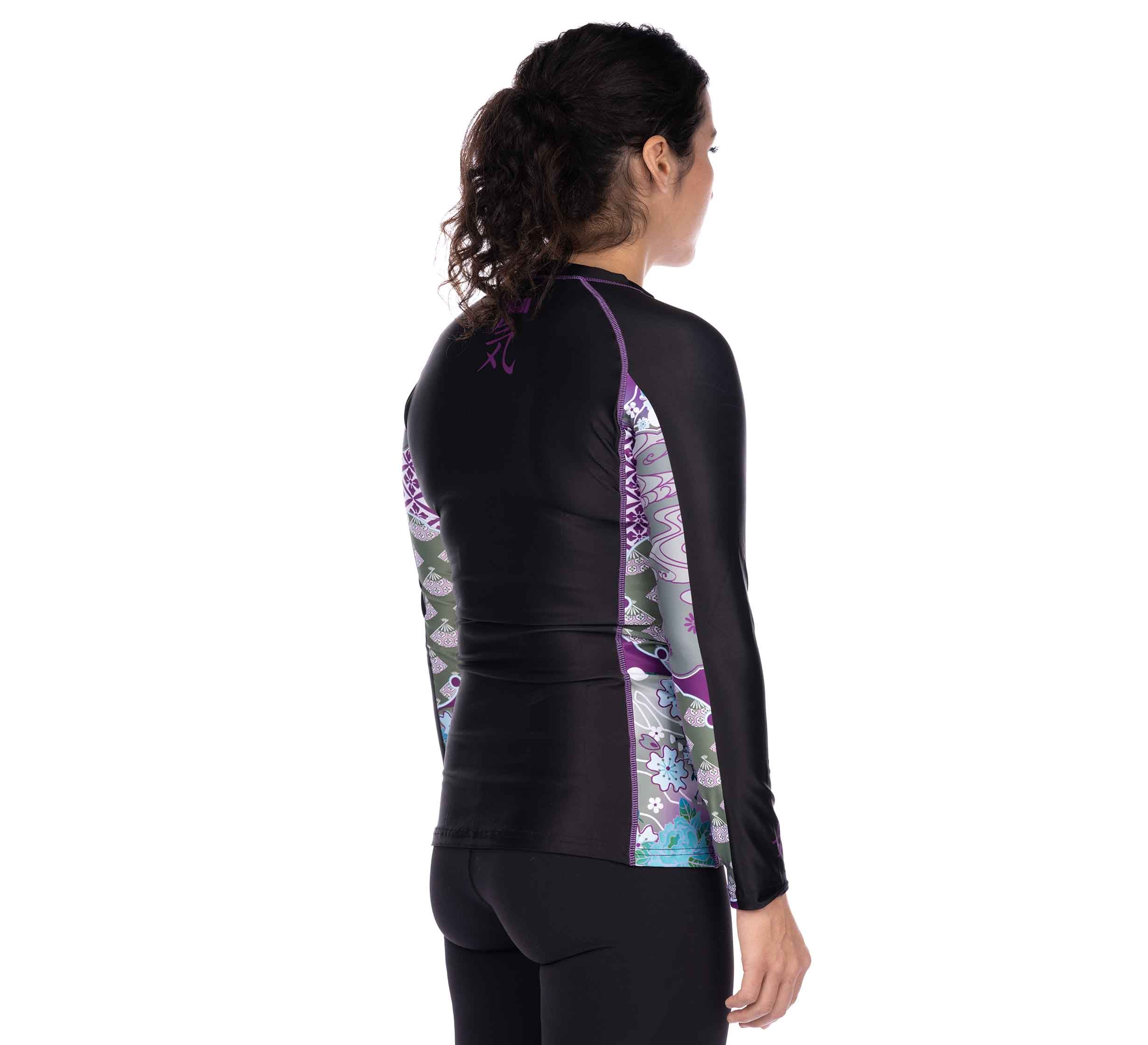 A woman with dark curly hair, wearing the Kimono Womens Long Sleeve Rashguard Purple and black leggings, stands facing away from the camera against a white background.