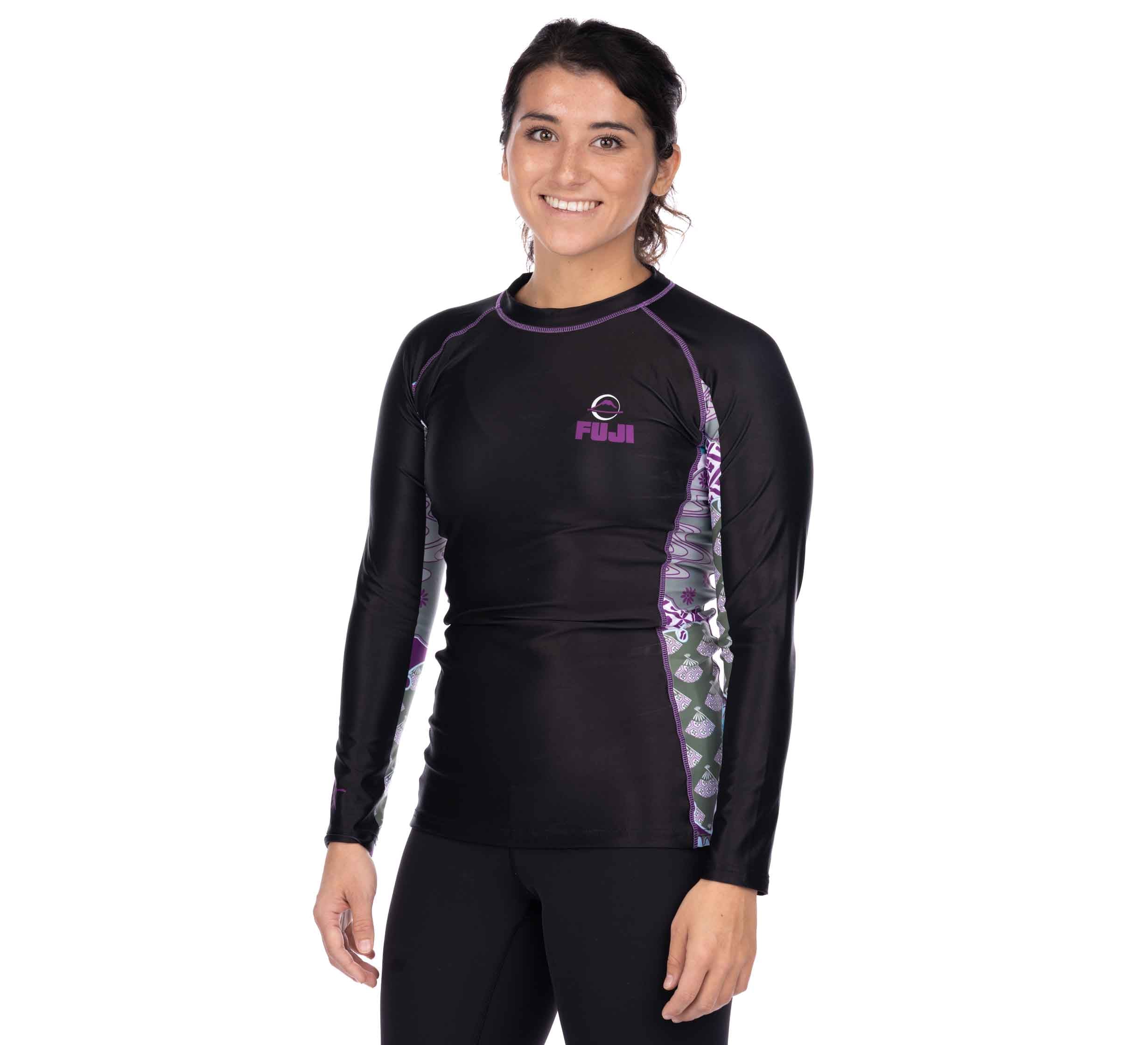 A woman wearing the Kimono Womens Long Sleeve Rashguard Purple, featuring black with purple and patterned side panels, stands and smiles against a plain white background.