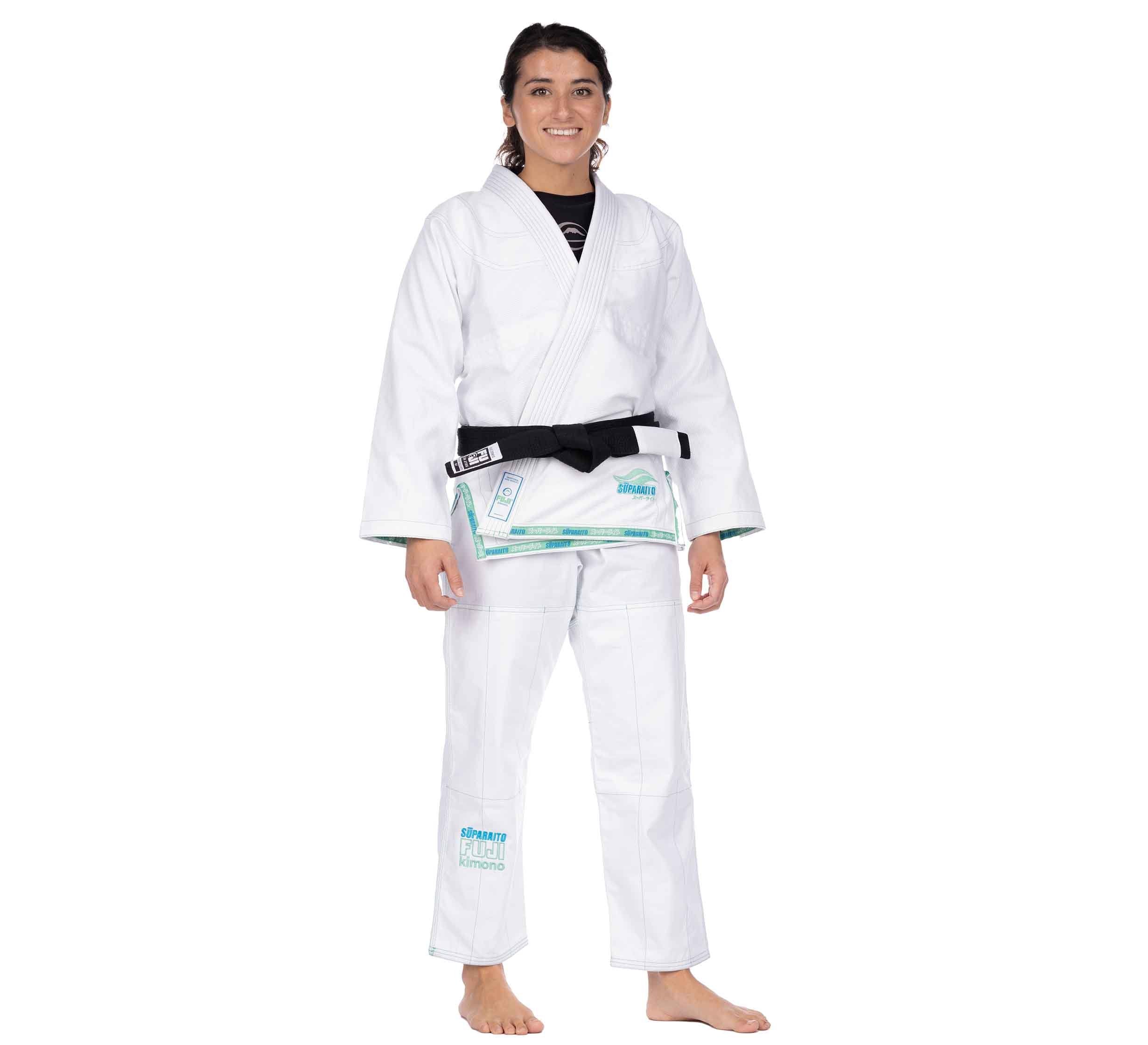 A woman stands barefoot and smiling in the Suparaito Girl's BJJ Gi Teal, featuring a black belt and blue-turquoise patches, against a plain white background.
