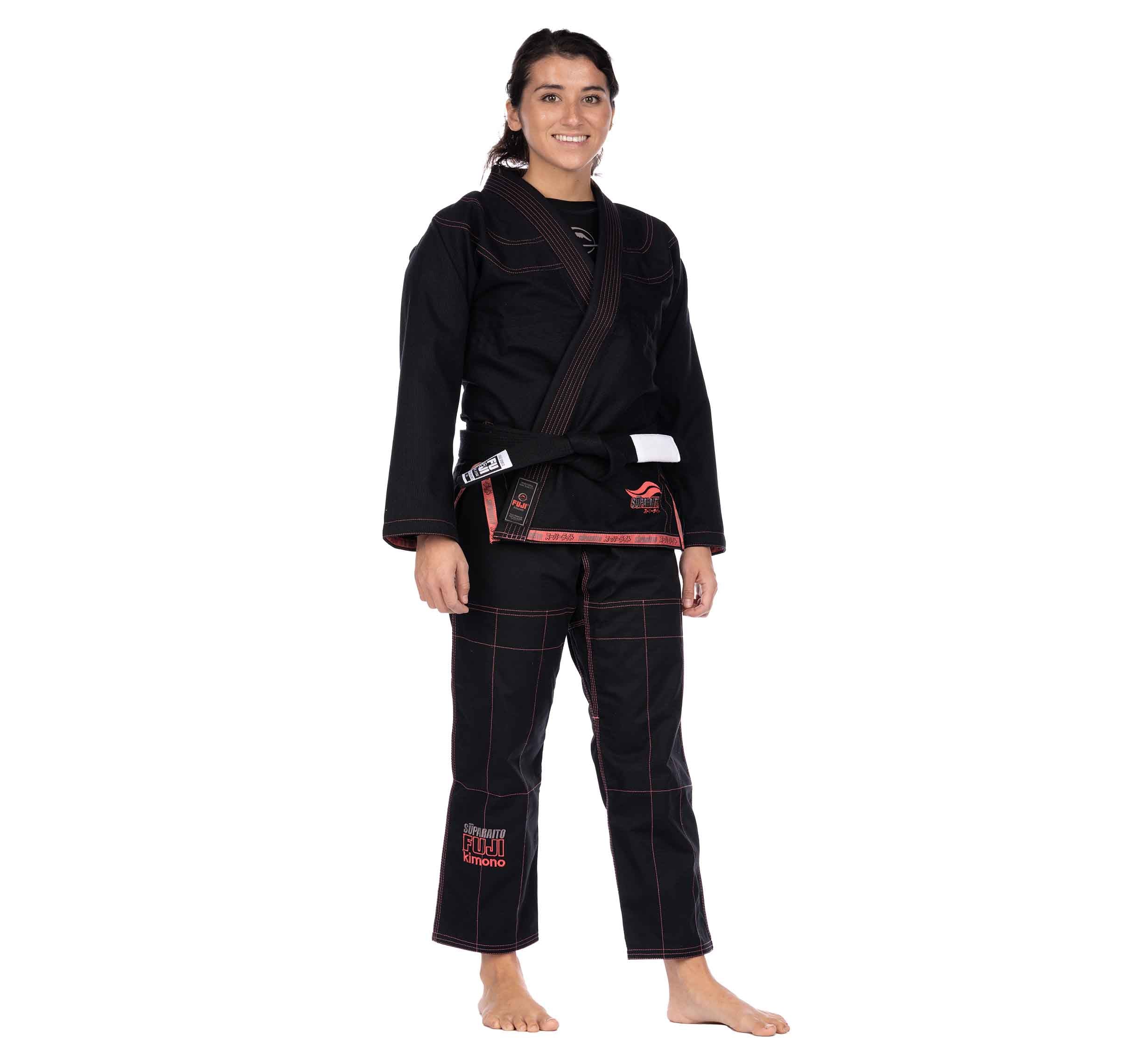 A woman wearing the Suparaito Womens BJJ Gi in pink with a brown belt stands barefoot and smiling, facing the camera against a plain white background.