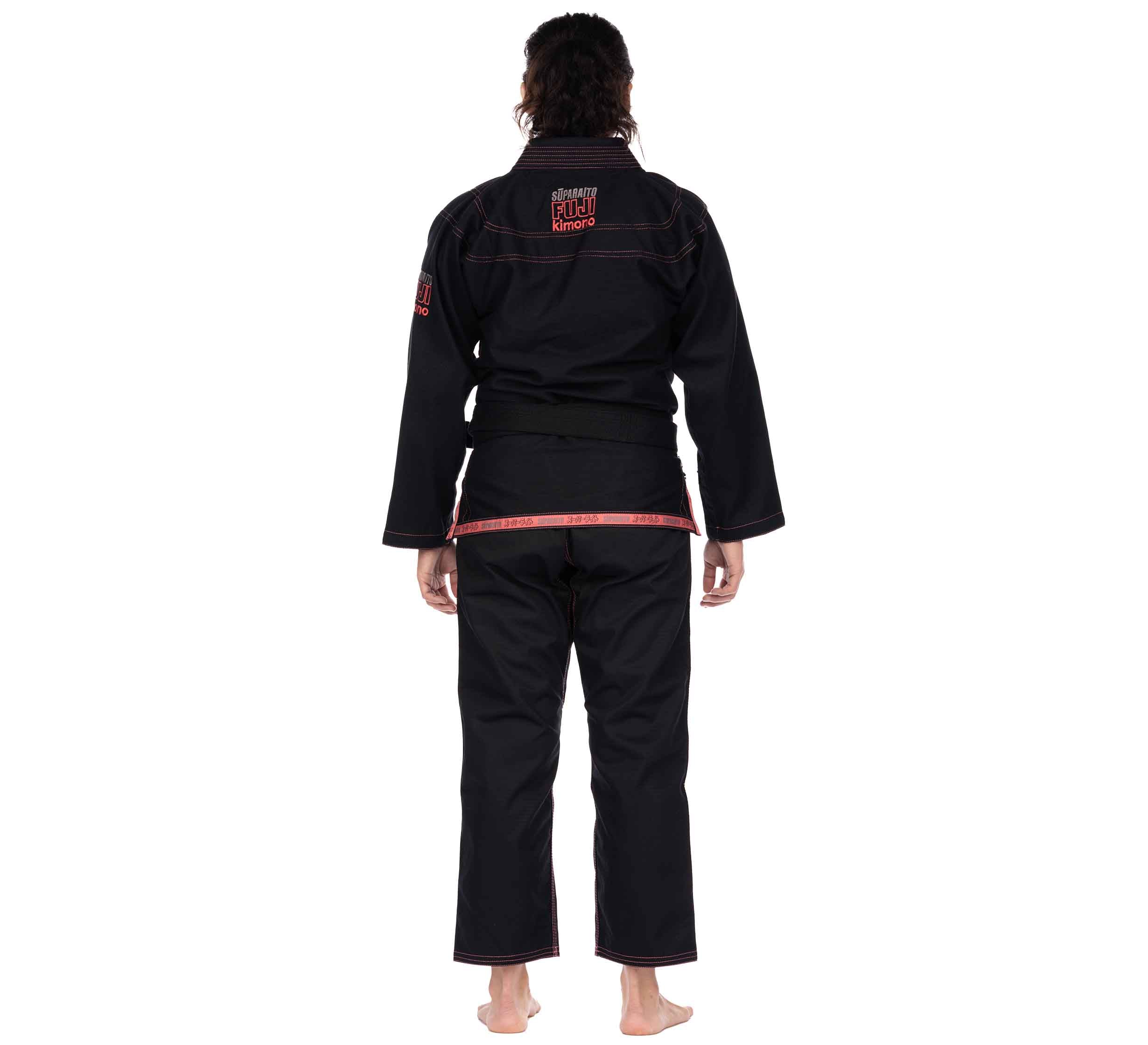A person stands barefoot facing away, wearing the Suparaito Womens BJJ Gi in pink with a black belt. The gi features embroidered text and patches on the back. The background is plain white.