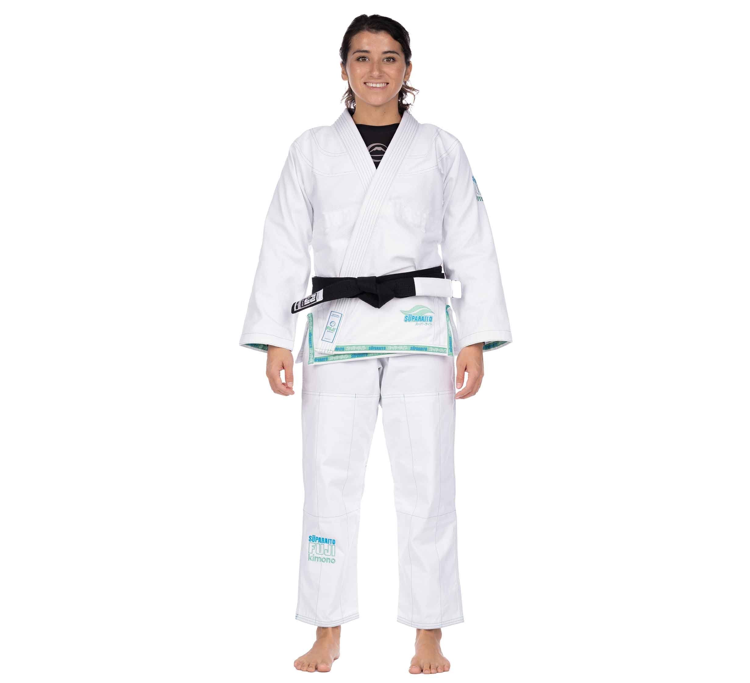 A barefoot, smiling person wears the Suparaito Girl's BJJ Gi Teal with a black belt. The white gi features teal accents and embroidered patches, shown against a plain white background.