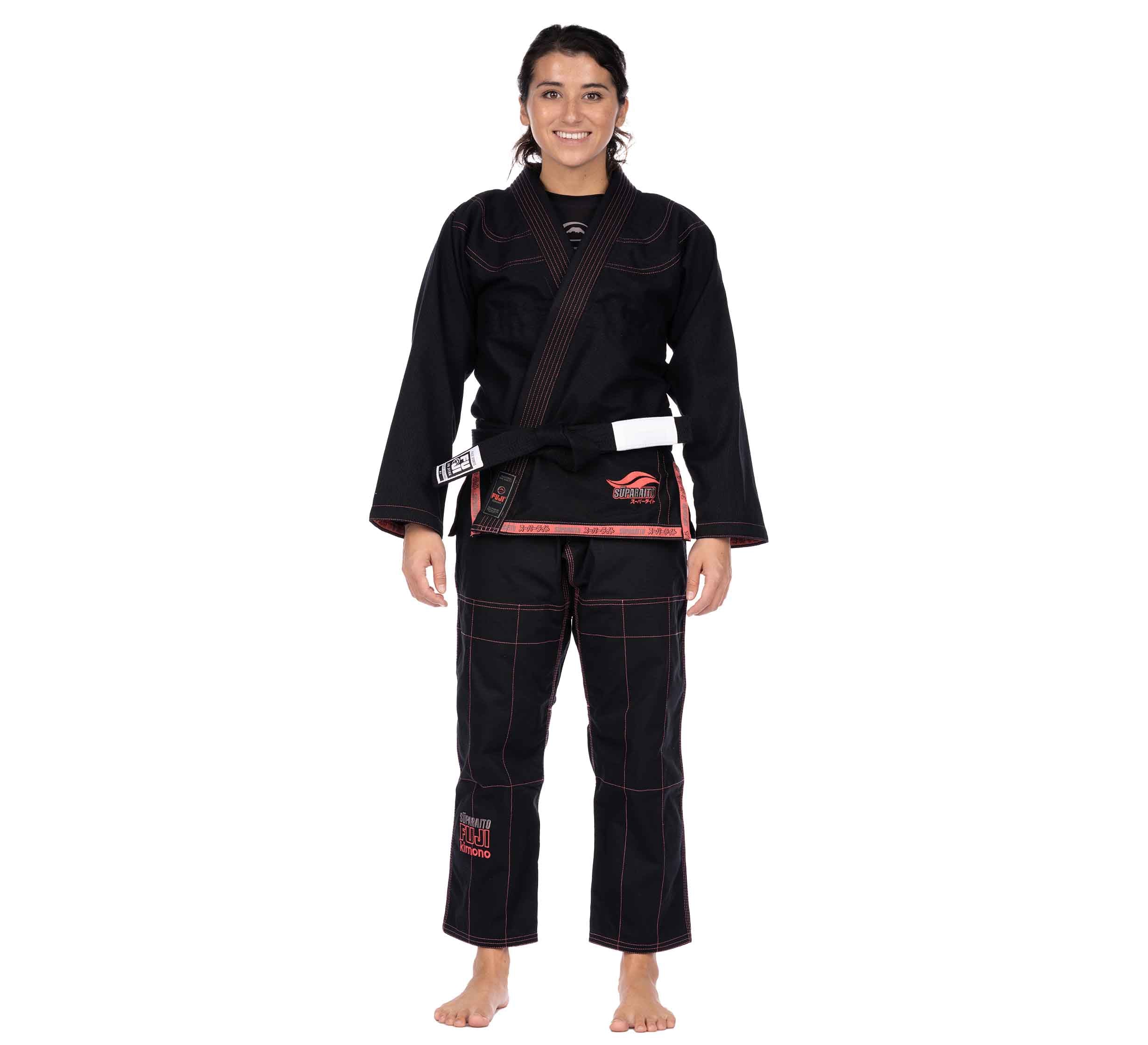 A woman smiles while standing barefoot in a Suparaito Womens BJJ Gi Pink with a brown belt against a white background.