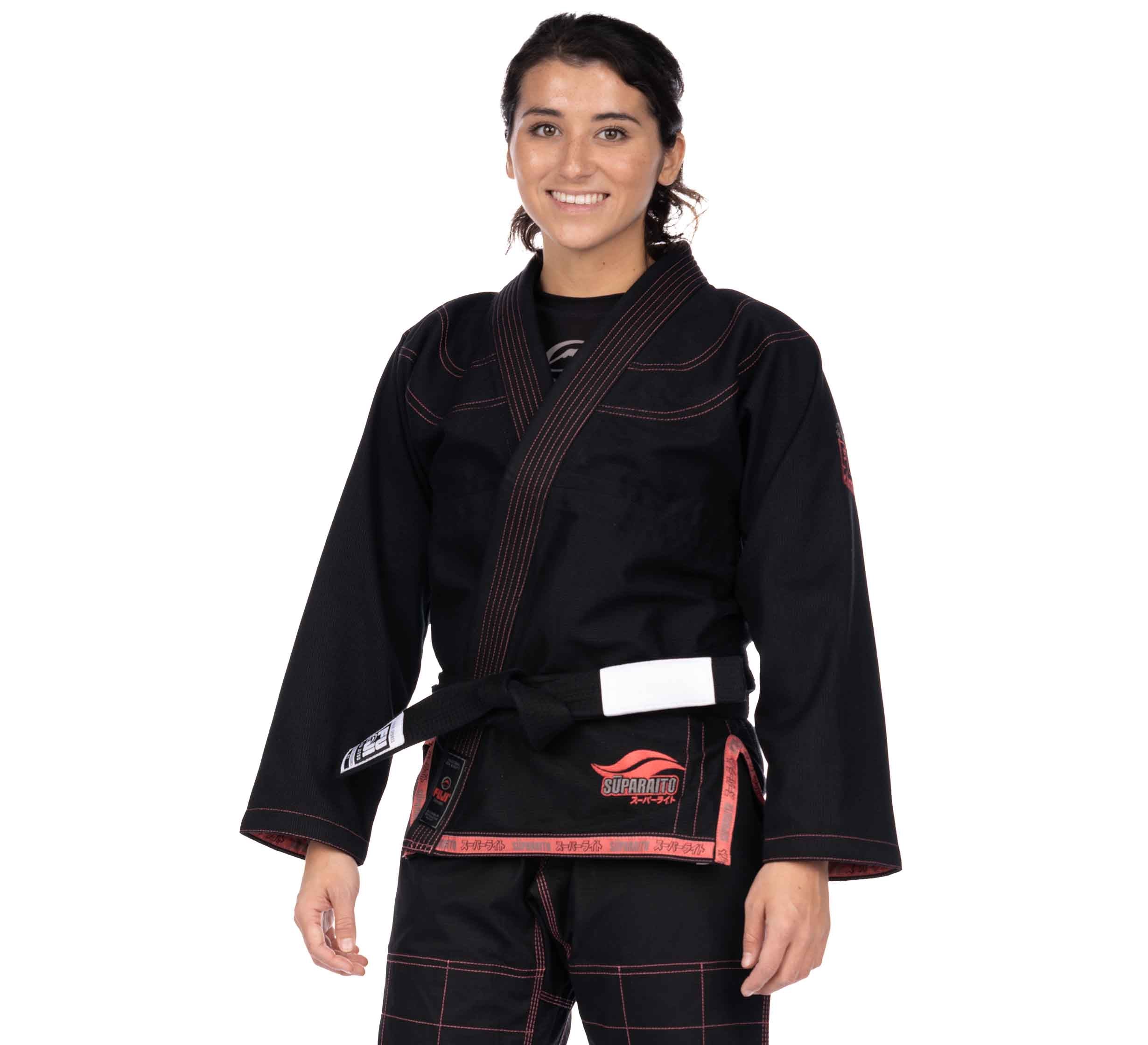 A person wearing the Suparaito Womens BJJ Gi Pink with patches and logos on the chest and waist, along with a white belt, stands facing the camera and smiling. The background is plain white.