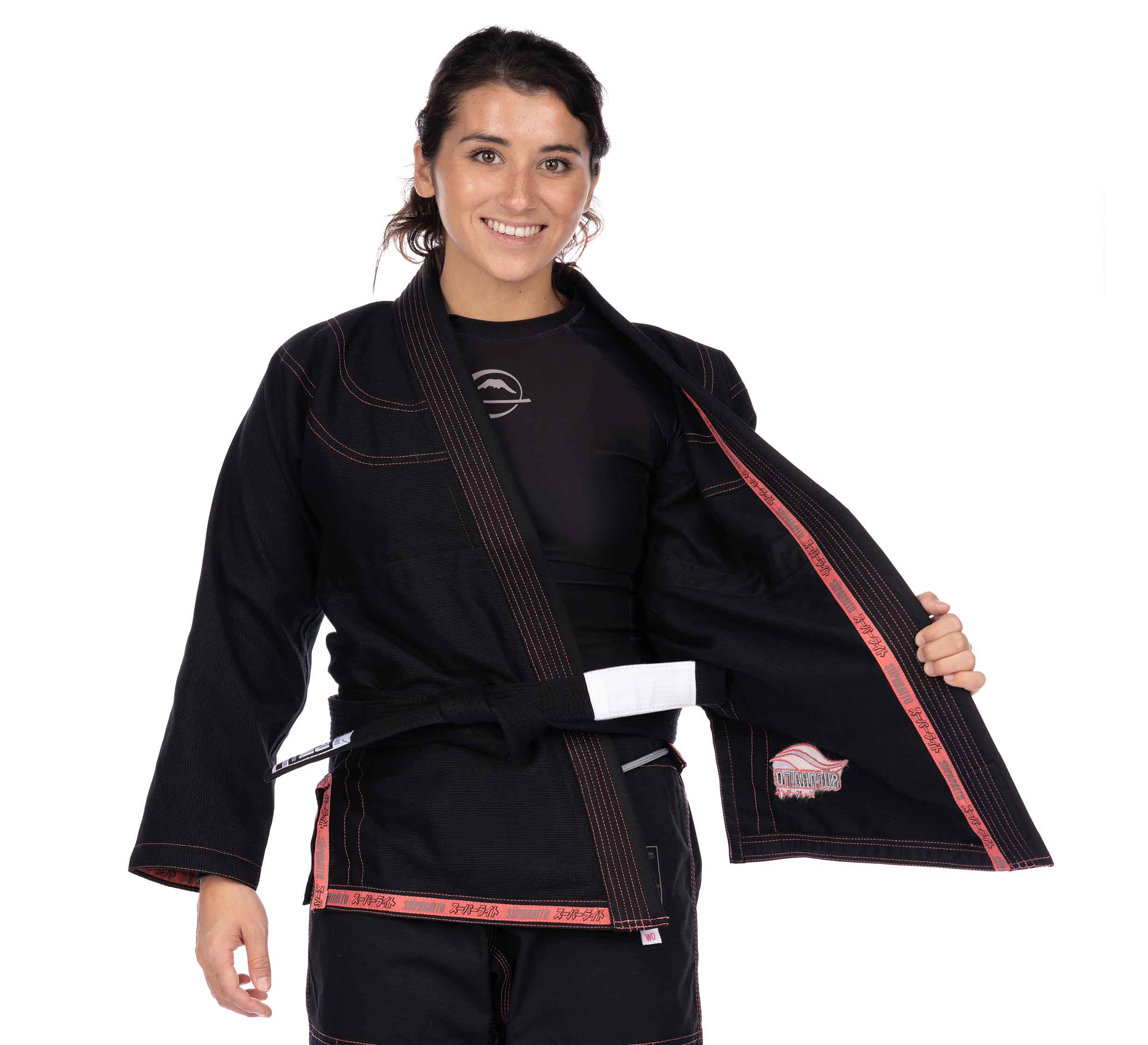 A woman smiles while wearing the Suparaito Womens BJJ Gi Pink with a black belt, holding open the jacket to show the inside. She stands against a plain white background.