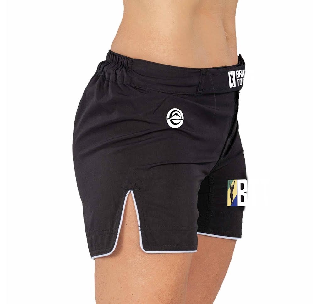 A person wears BTT Womens Baseline Shorts Black, featuring white trim, front slit, small white logos, and an elastic waistband. Only their midsection and legs are shown.