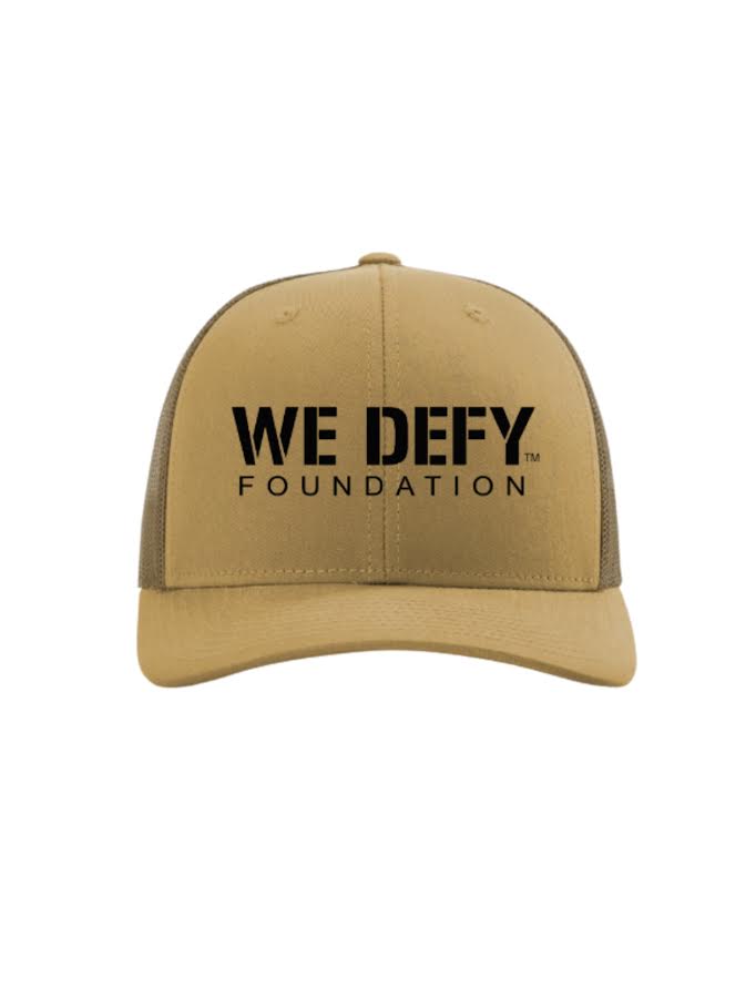 The We Defy Trucker Hat Amber Gold is a tan and brown hat with "WE DEFY FOUNDATION" in bold black letters on the front panel.