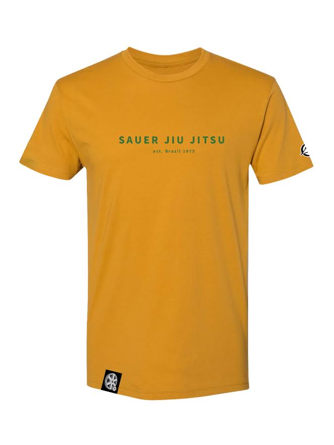 Introducing the Pedro Sauer Jiu Jitsu T-Shirt in Antique Gold, featuring "Sauer Jiu Jitsu est. Brazil 1975" printed in green across the chest, with small black and white patches on the left sleeve and bottom front hem.