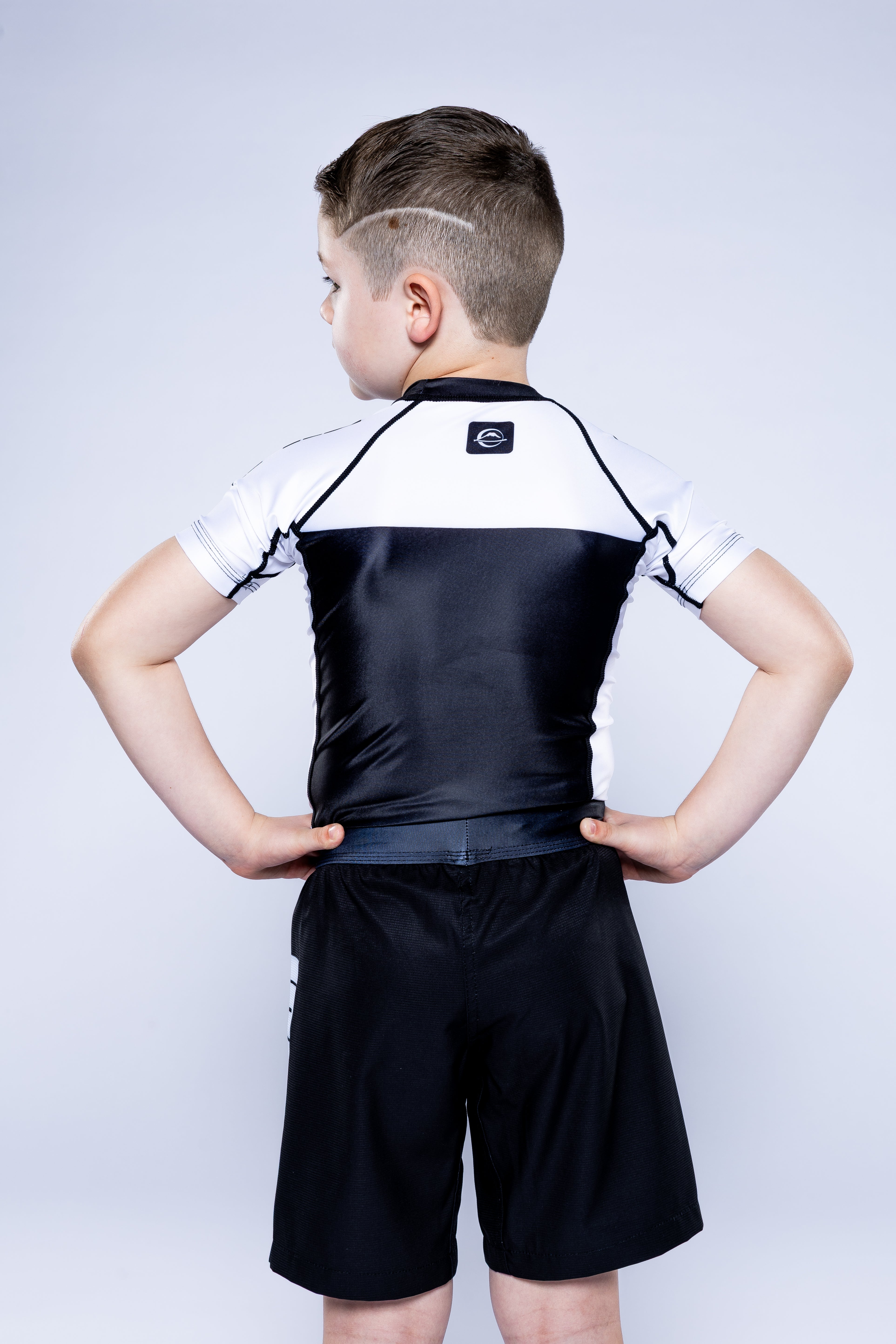 A young boy stands with hands on hips, wearing the Kids Competition Ranked Rashguard White and black shorts, against a plain light background.