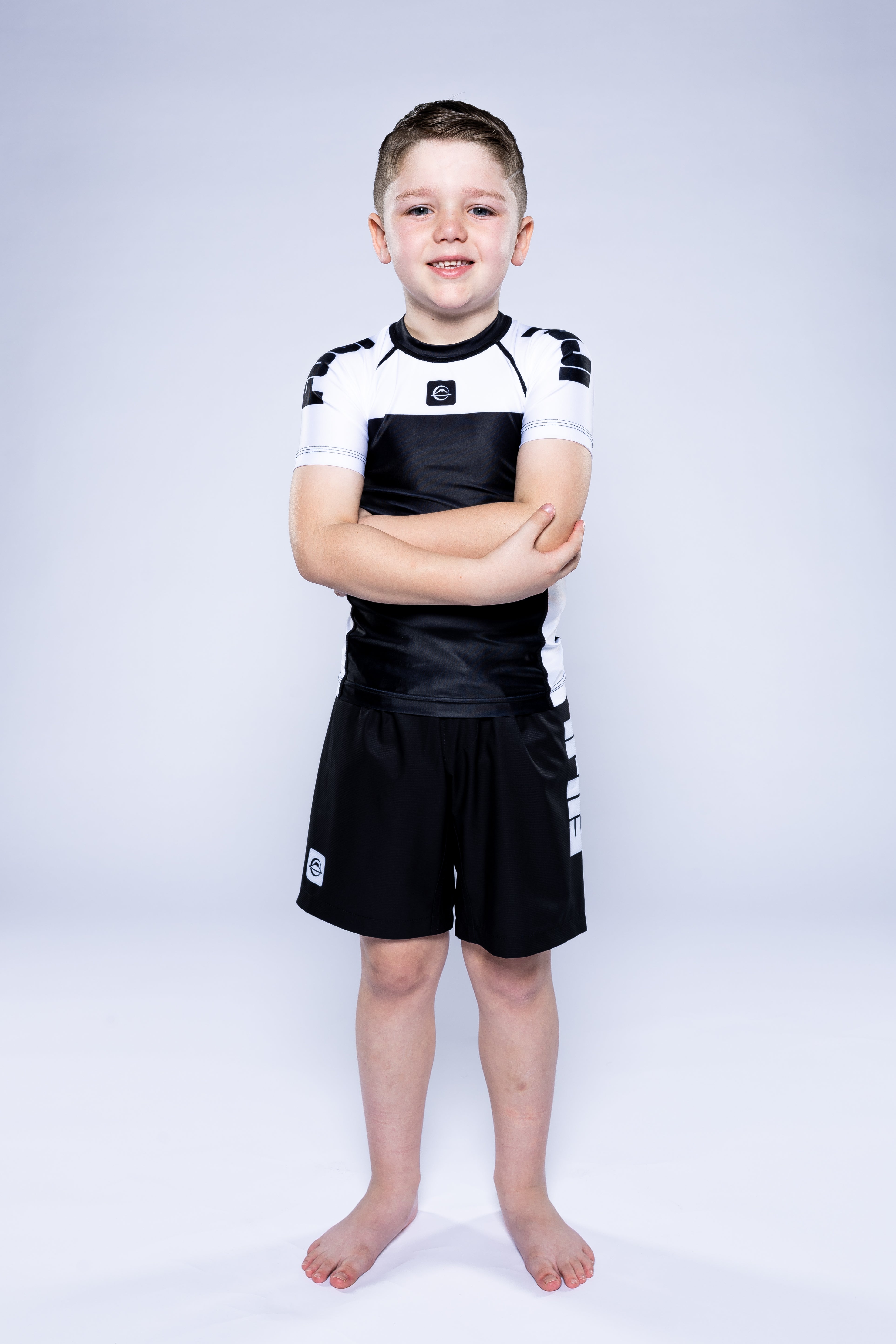 A young boy stands barefoot with arms crossed, smiling, wearing the Kids Competition Ranked Rashguard White with matching shorts against a plain white background.