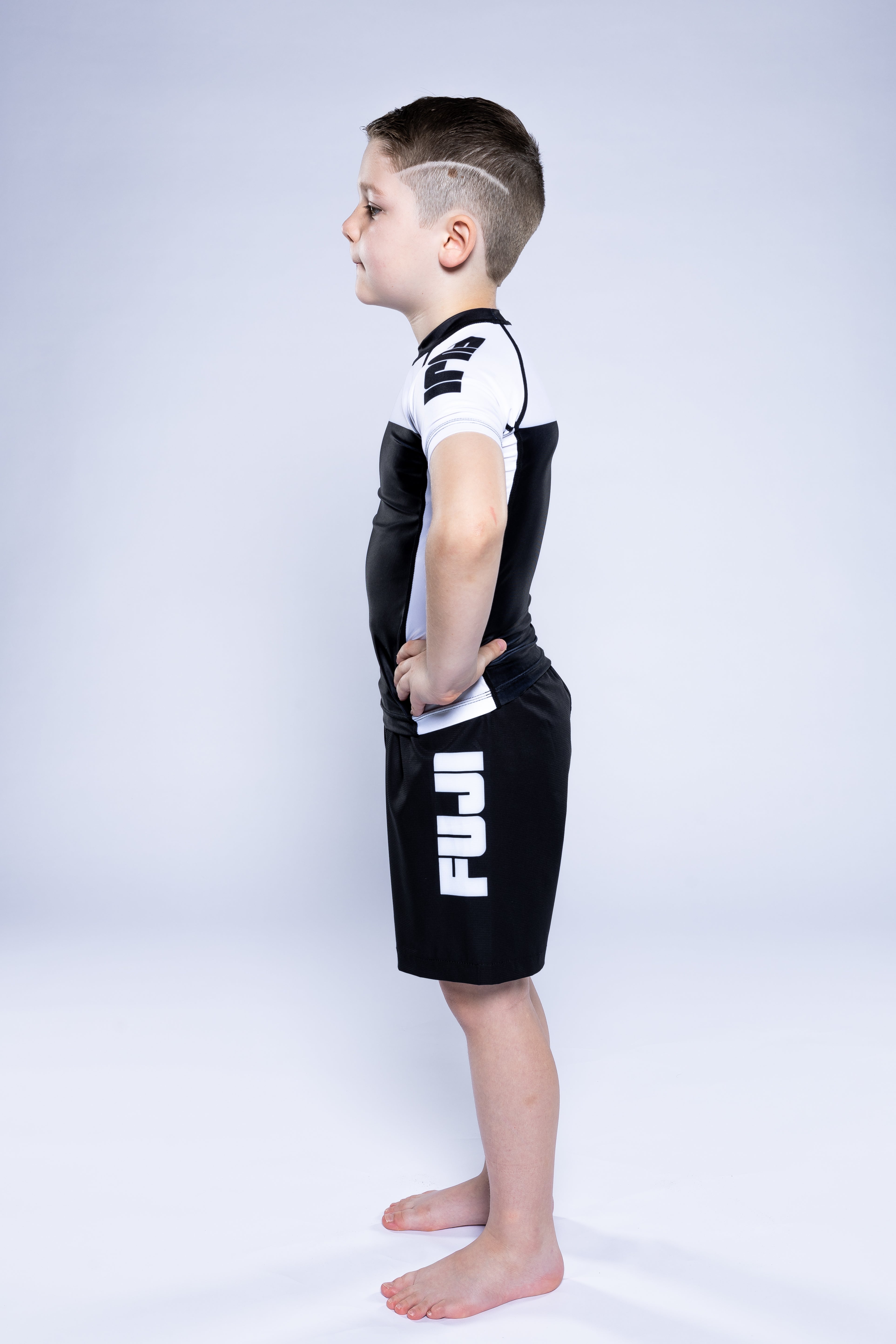 A young boy stands barefoot in profile on a white background, wearing the Kids Competition Ranked Rashguard White and matching black and white FUJI shorts, with his hands on his hips.