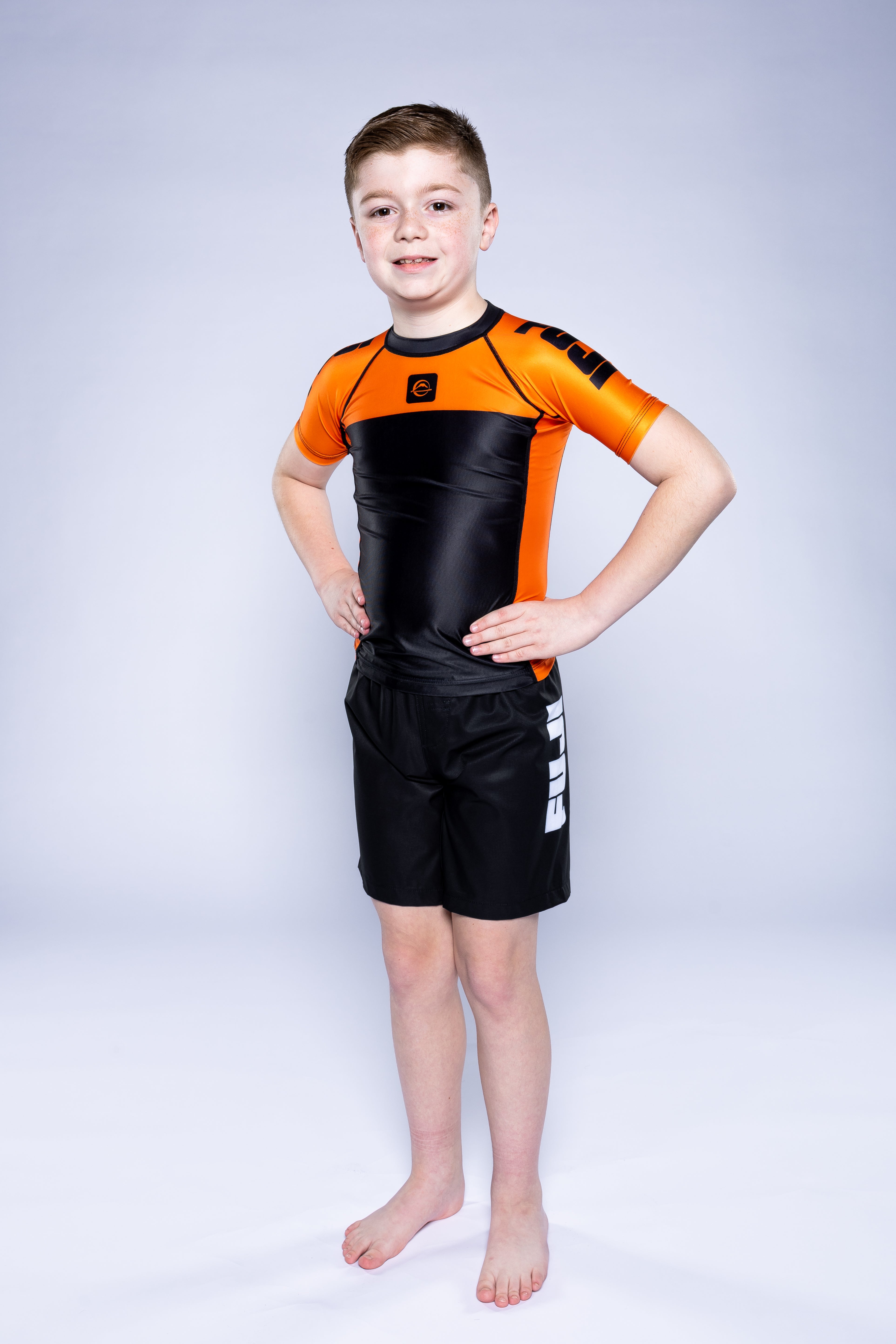 A young boy stands barefoot with hands on hips, confidently posing in the Kids Competition Ranked Rashguard Orange and black shorts with white lettering against a light gray background.
