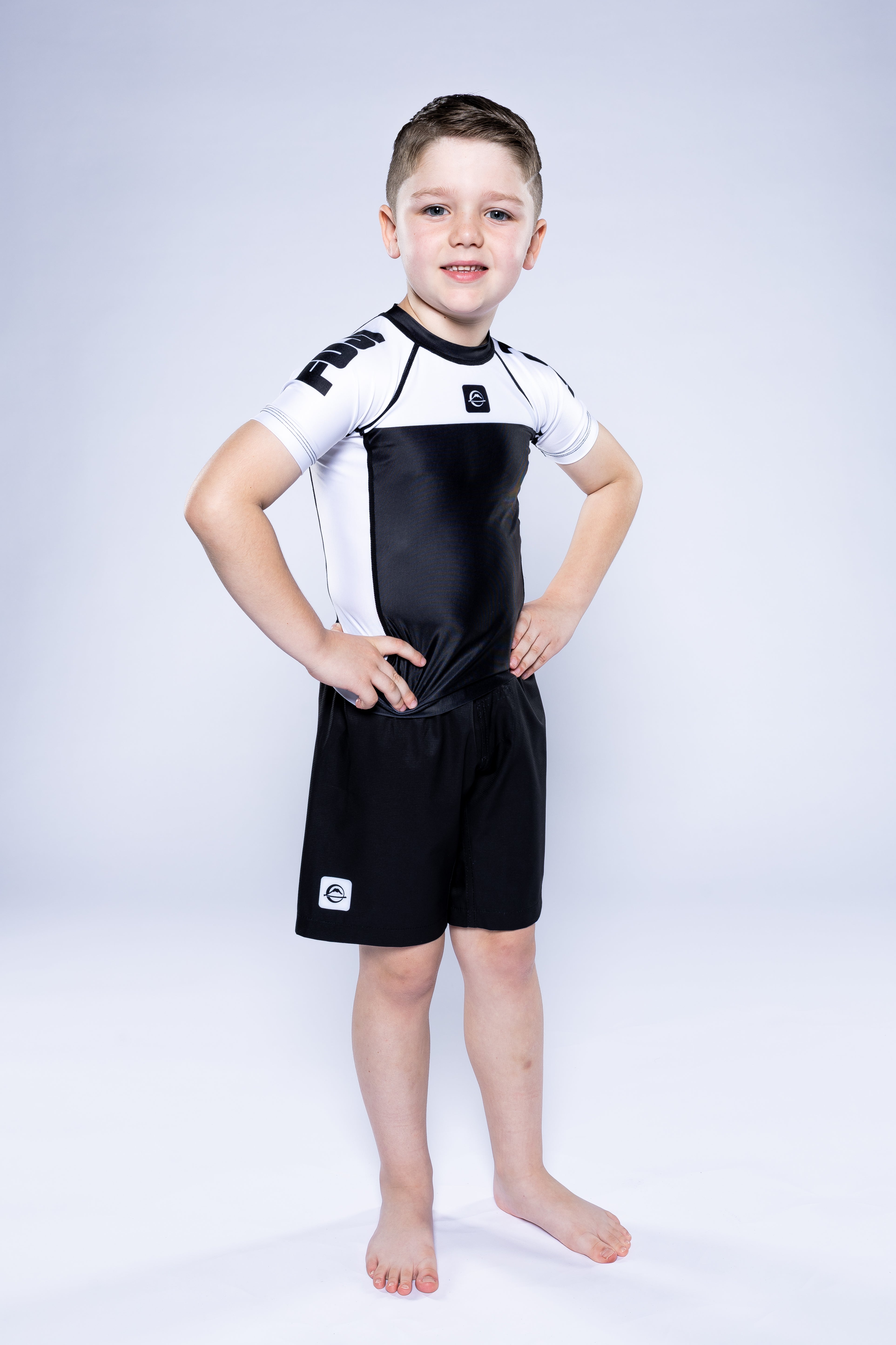 A young boy stands barefoot on a white background, smiling confidently with hands on hips and wearing the Kids Competition Ranked Rashguard White with matching shorts.