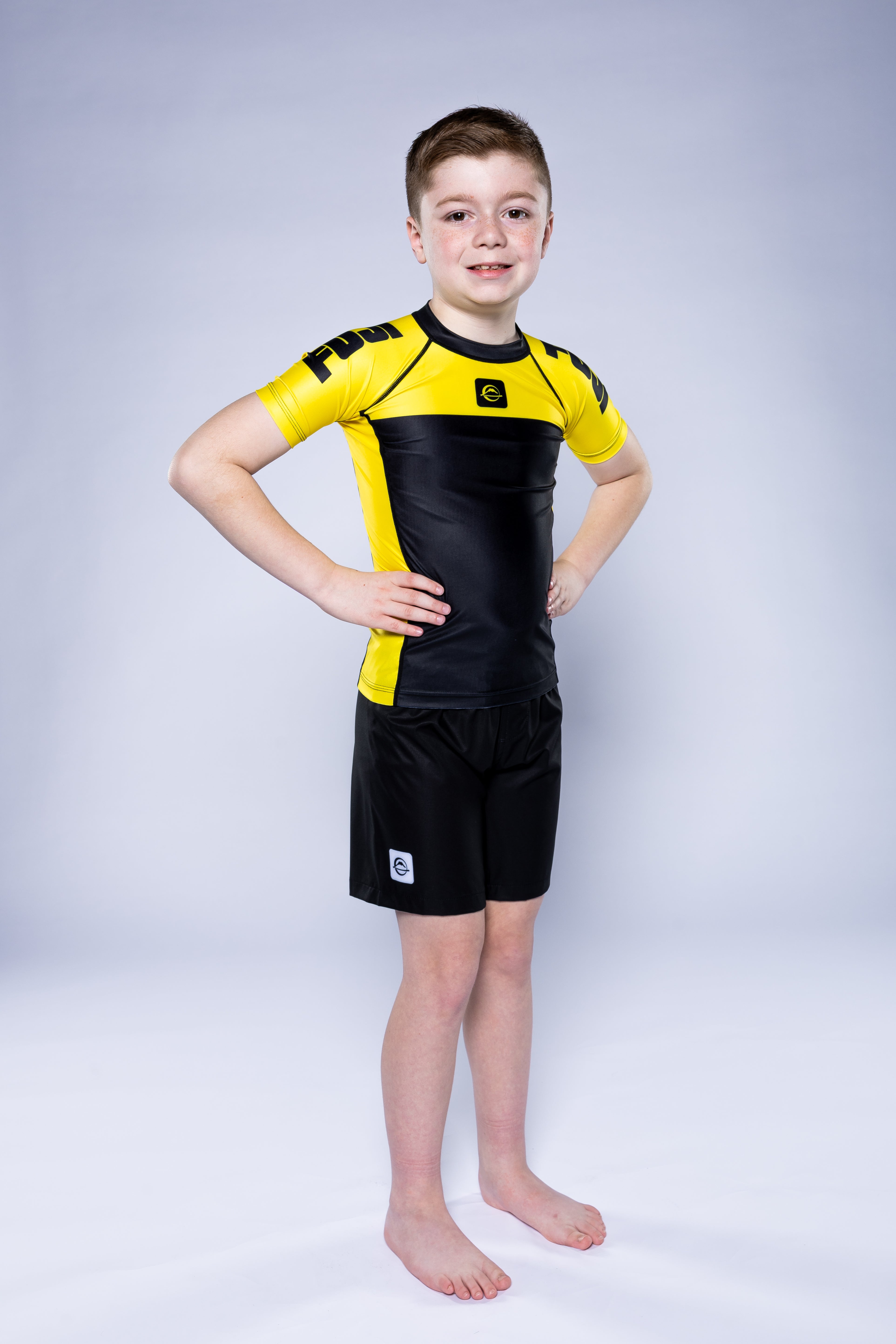 A young boy stands barefoot on a white background, smiling with his hands on his hips while wearing the Kids Competition Ranked Rashguard Yellow.