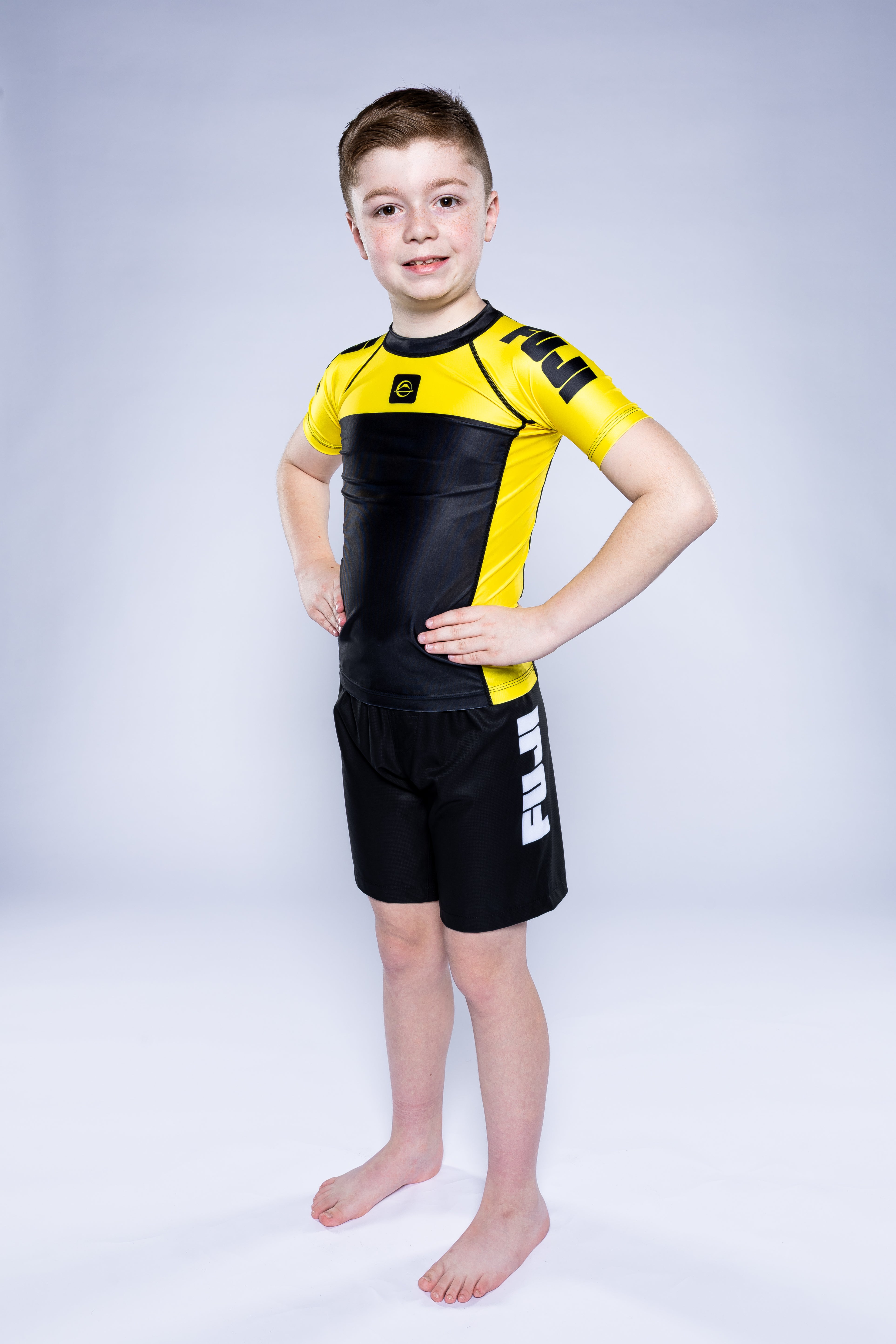 A young boy stands barefoot on a white background, smiling confidently with his hands on his hips. He is wearing the Kids Competition Ranked Rashguard Yellow and black shorts featuring the FUJI logo on the side.
