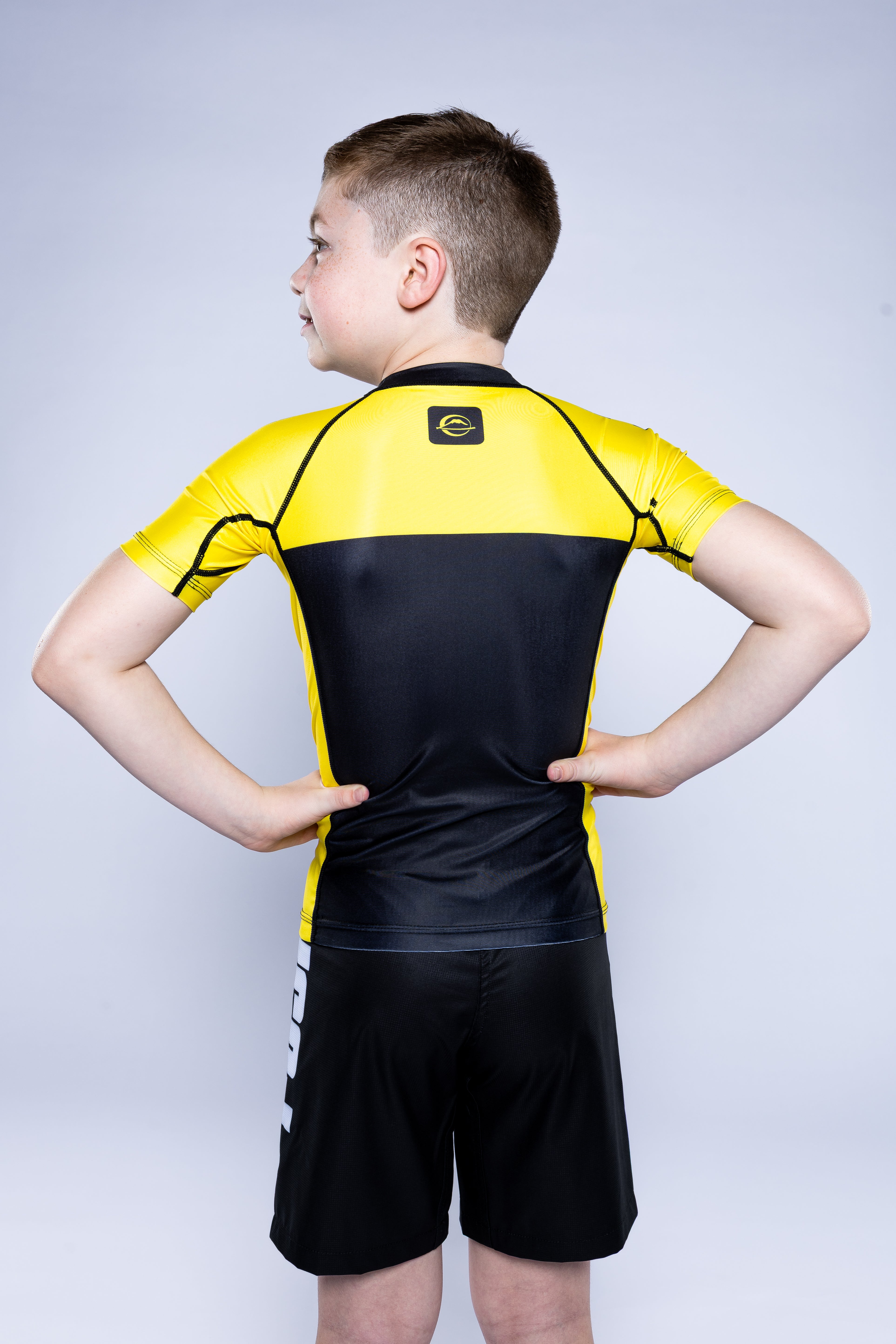 A boy stands with his hands on his hips, facing away from the camera, wearing the Kids Competition Ranked Rashguard Yellow and black shorts against a plain light background.