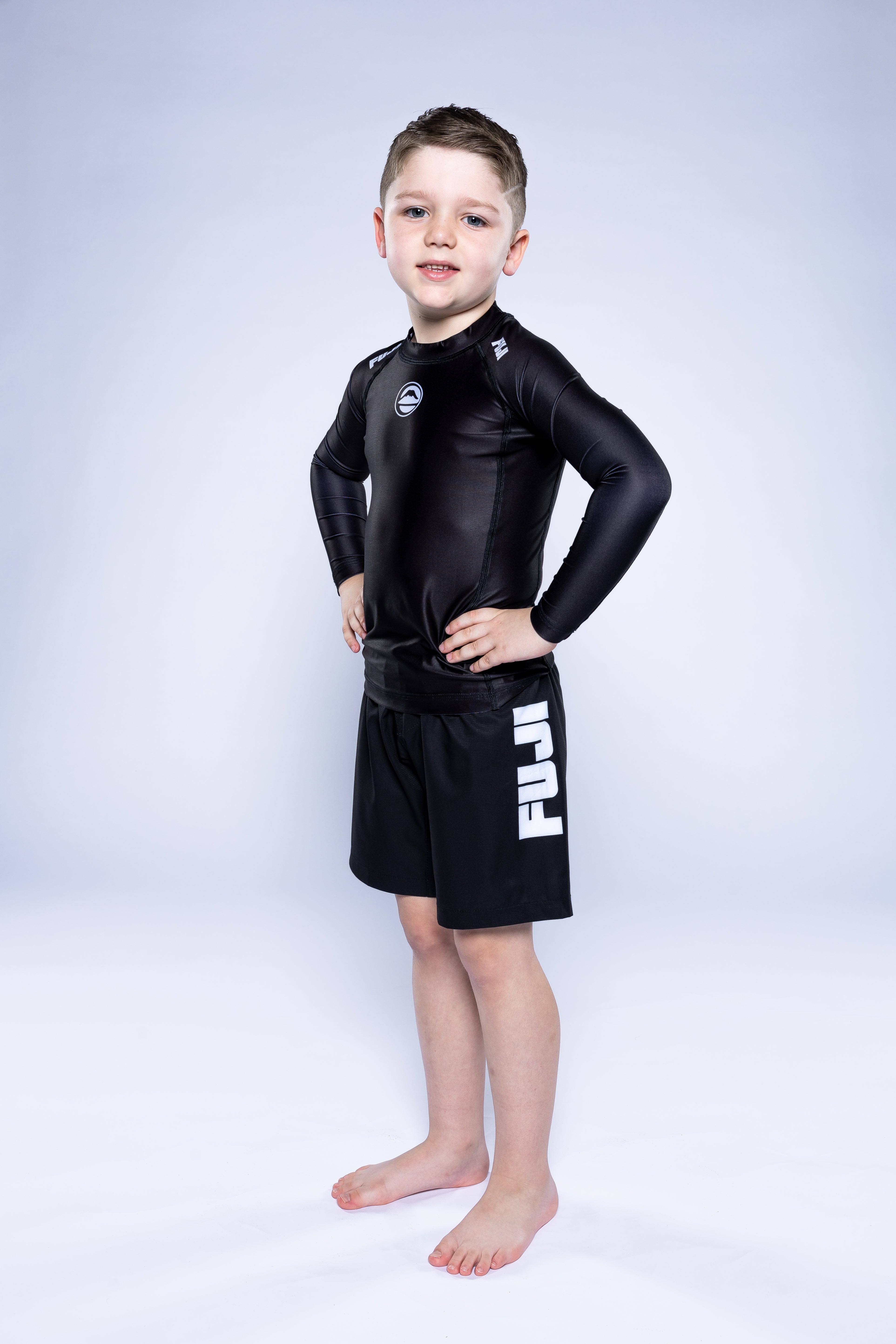 A young boy stands barefoot on a white background, confidently smiling with his hands on his hips while wearing the Kids Baseline 2.0 Long Sleeve Rashguard Black and matching shorts with FUJI printed on the side.