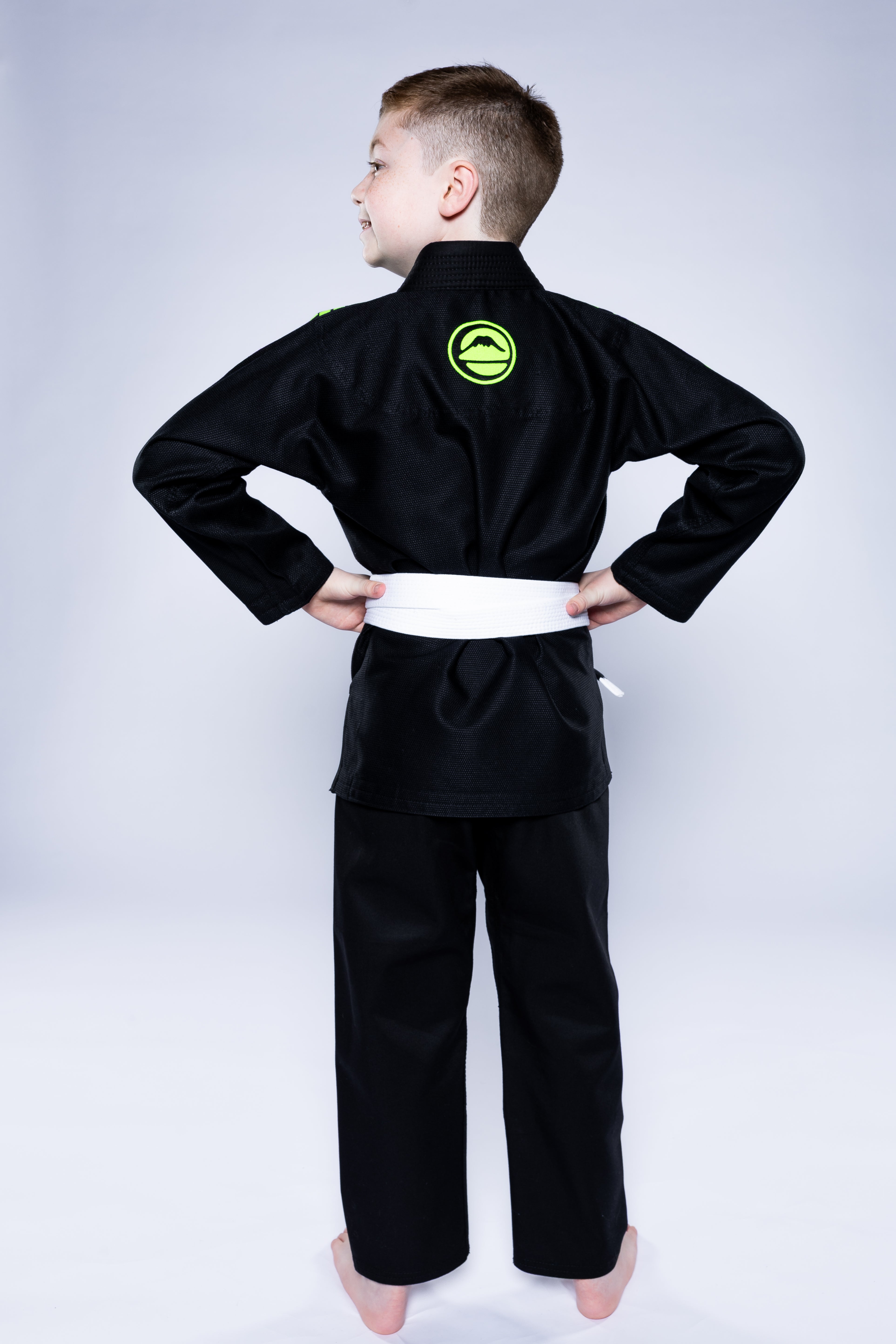 A young boy stands barefoot, hands on hips, facing away from the camera against a light background, wearing the Kids Baseline 2.0 Slim Fit BJJ Gi Black Green with a white belt.