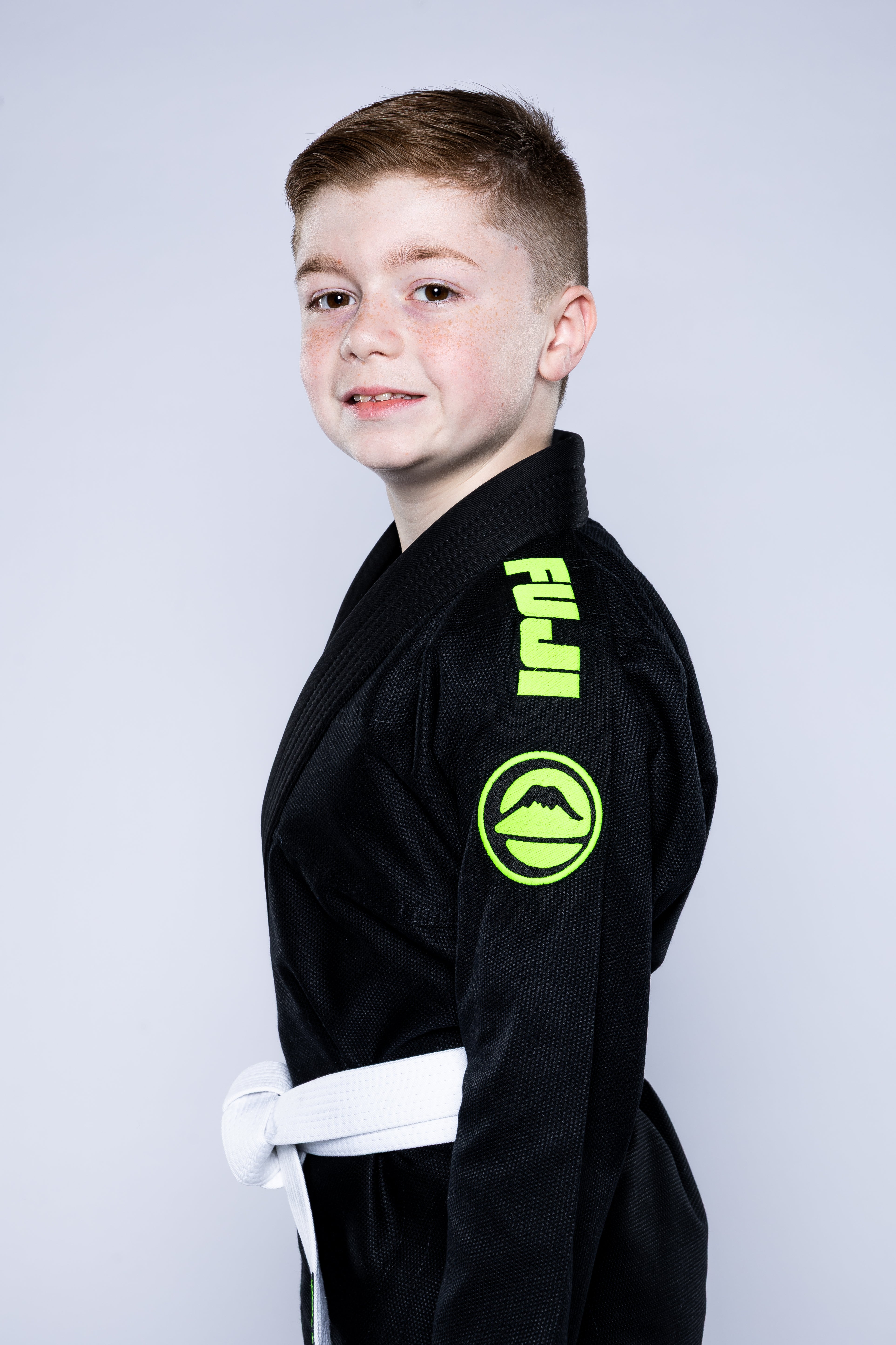 A young boy with short light brown hair smiles at the camera, wearing the Kids Baseline 2.0 Slim Fit BJJ Gi Black Green with a white belt, standing sideways against a plain light background.