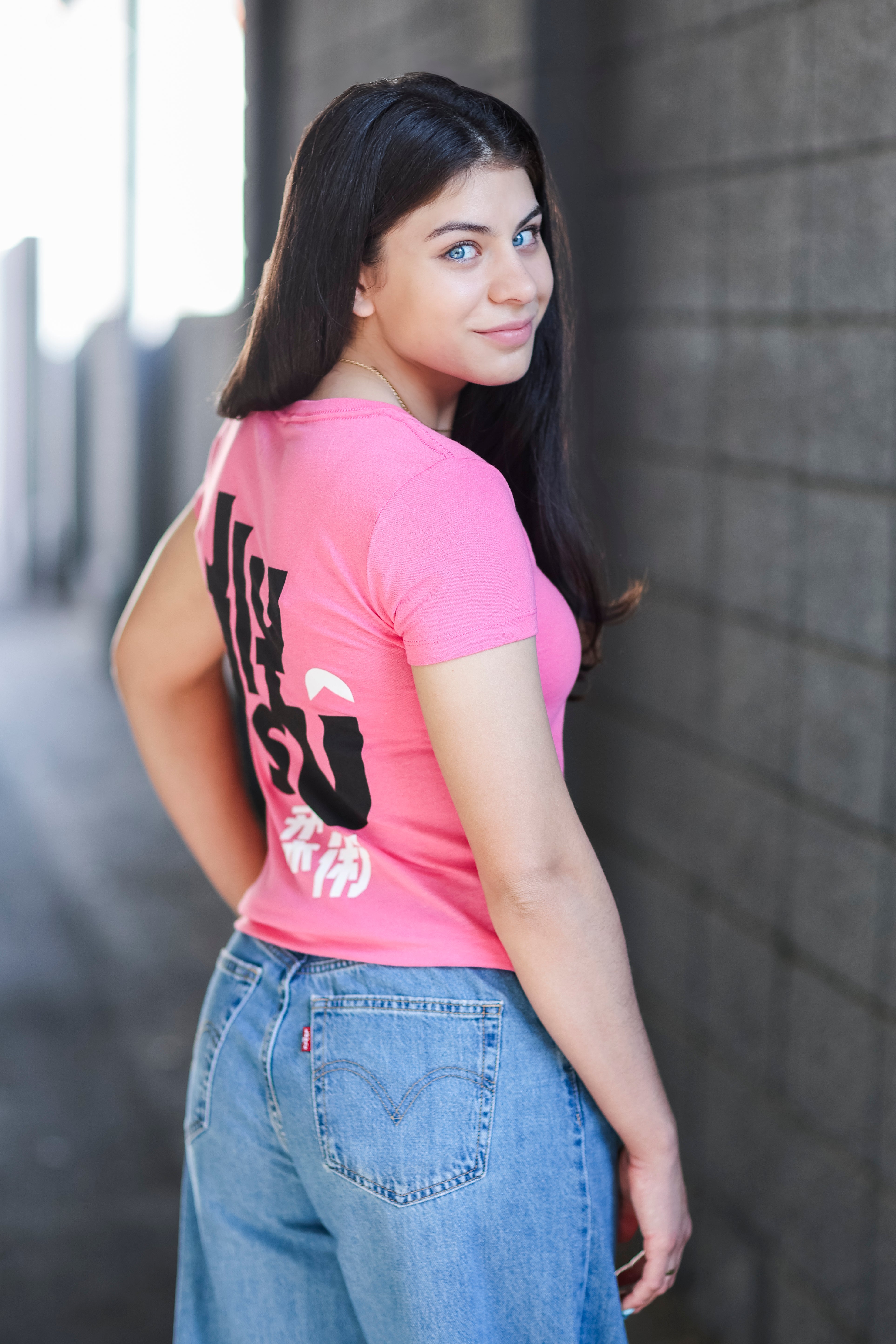 A young woman with long dark hair wearing a pink t-shirt with bold black and white text on the back and blue jeans, stands outdoors near a gray brick wall, looking over her shoulder and smiling softly.