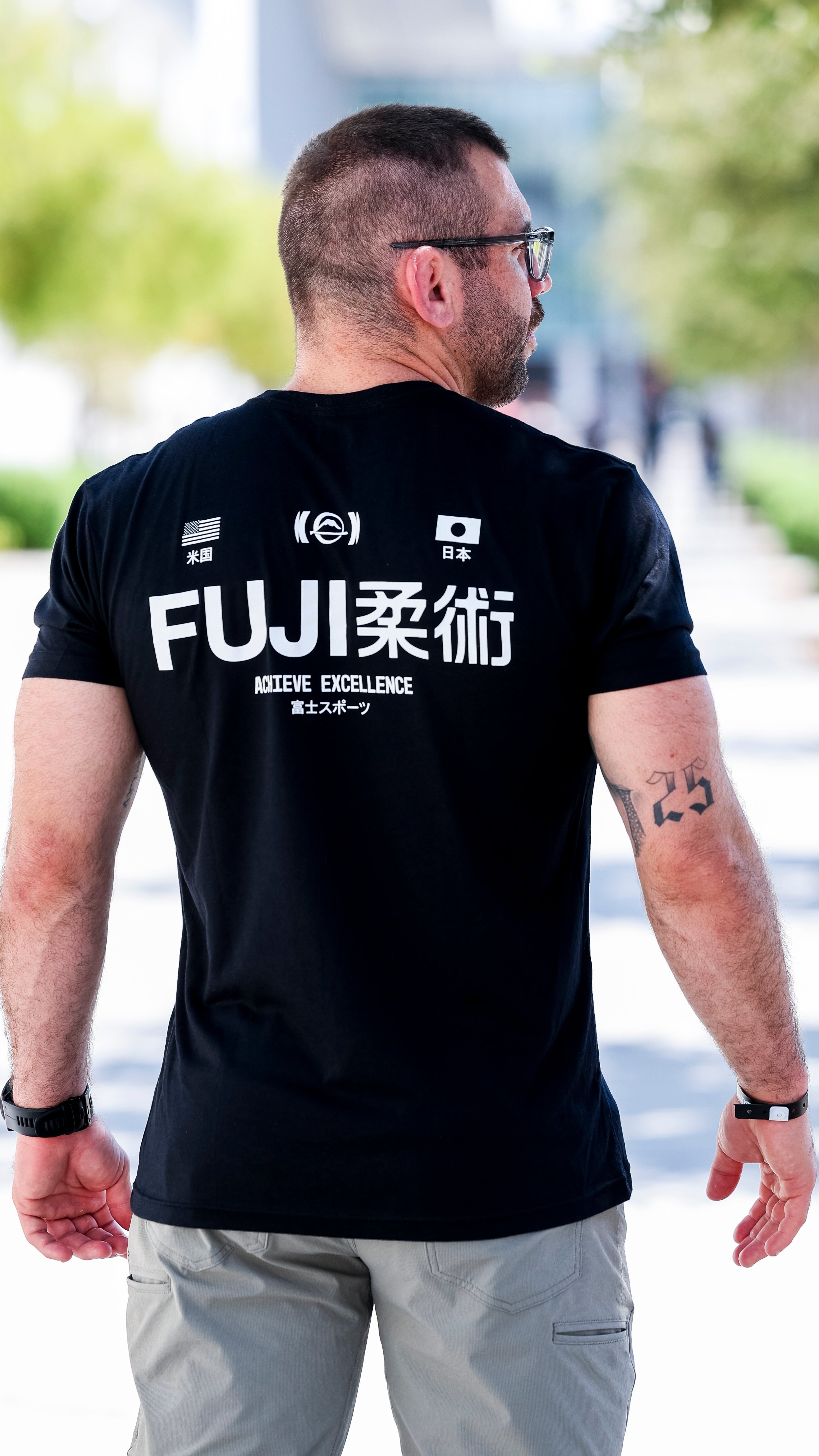 A man with short hair and a beard stands outdoors, facing away. He wears a black FUJI柔術 Achieve Excellence t-shirt and light gray pants. The number 25 is tattooed on his left arm. Trees and a walkway are visible ahead.