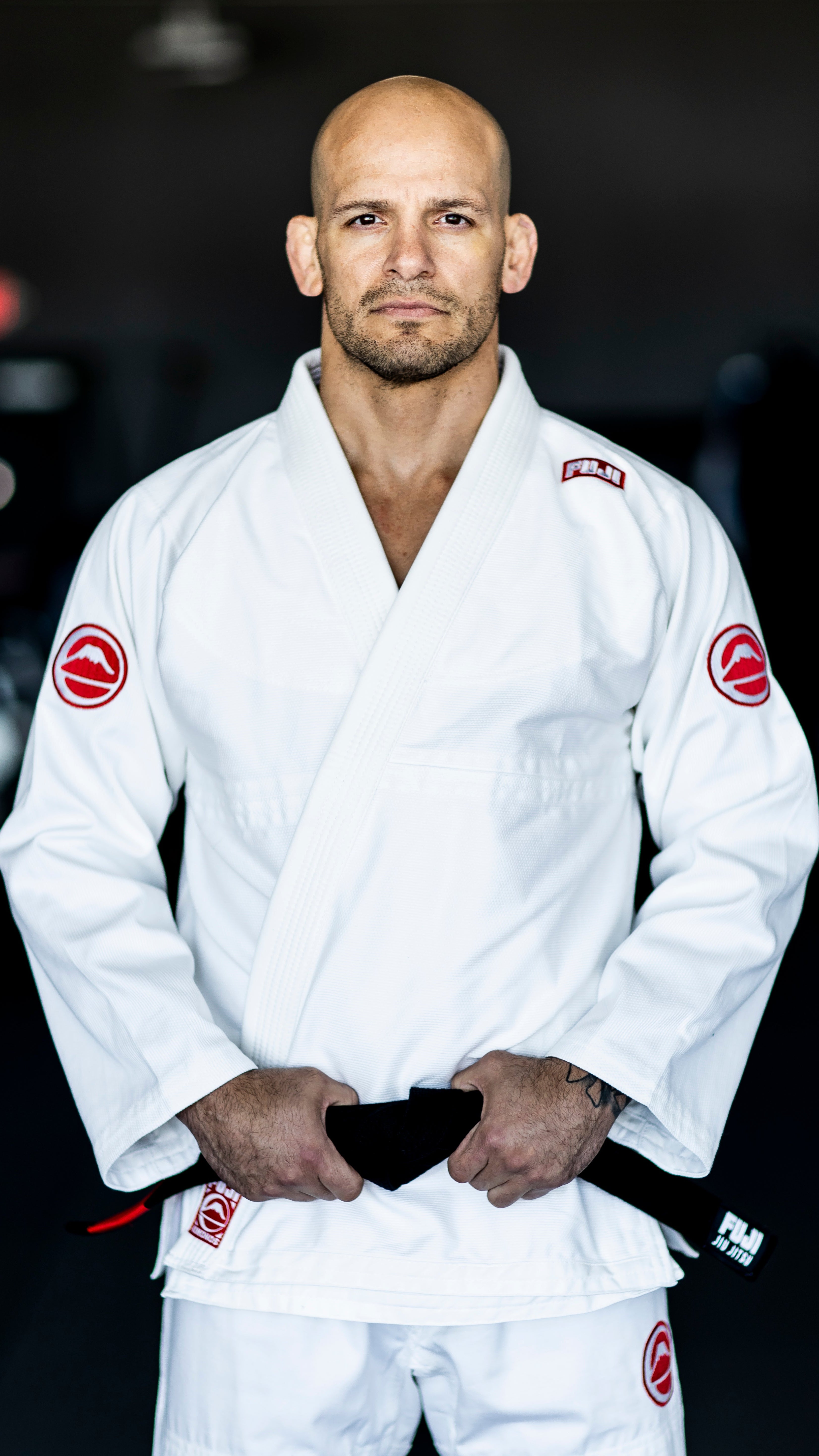 A person wearing a white Brazilian Jiu-Jitsu gi with red patches stands facing forward, holding a black belt with both hands. The background is dark and out of focus.