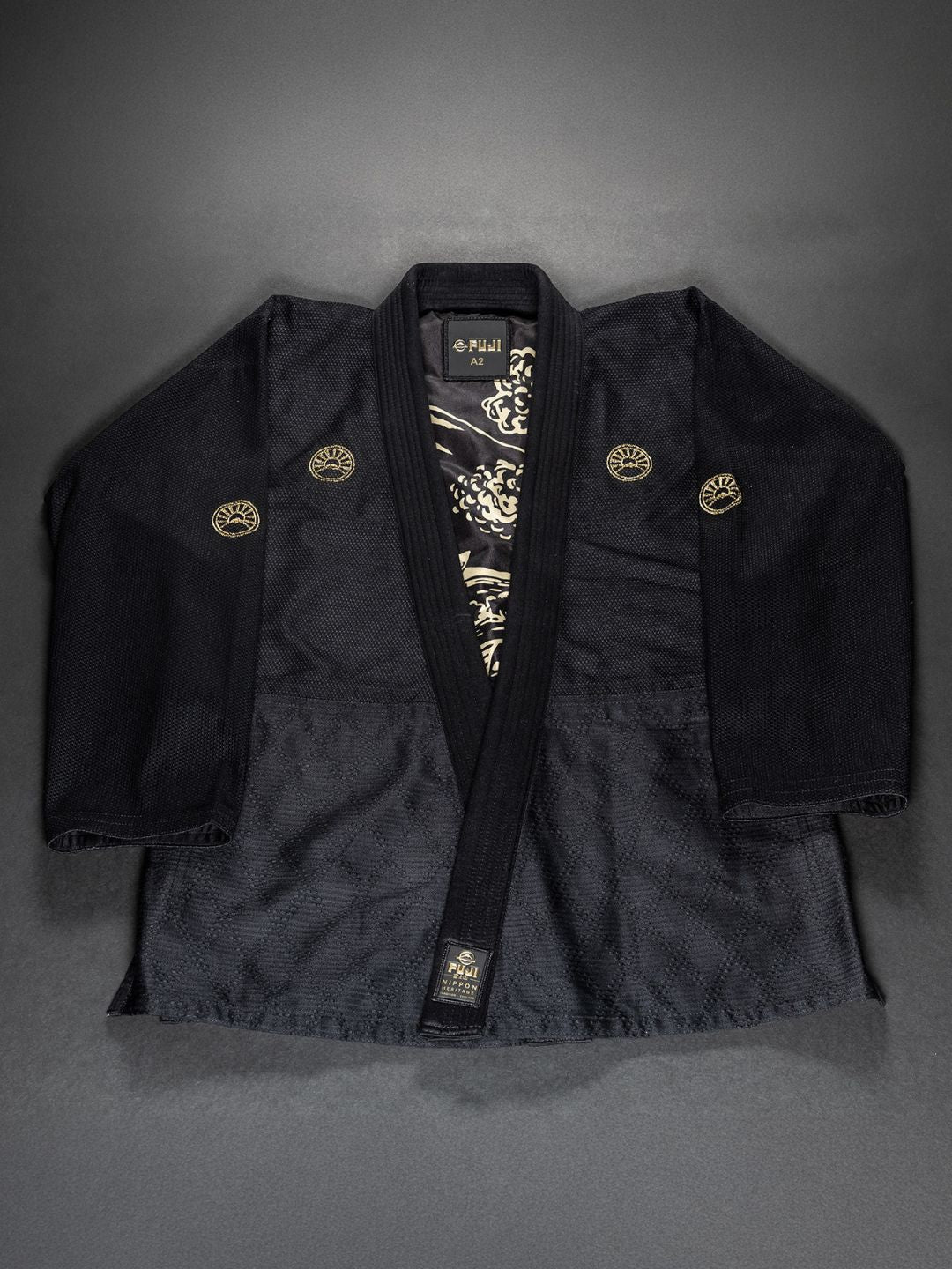 The Womens Heritage Keisho Lightweight BJJ Gi in black, featuring intricate gold embroidery and a dragon design on the inner lining, is displayed flat on a dark surface.