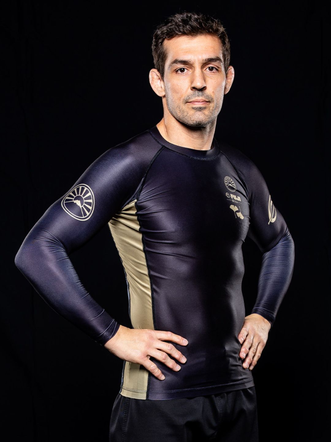 A man stands confidently with his hands on his hips against a black background, wearing the Heritage Keisho Long Sleeve Rashguard in black.