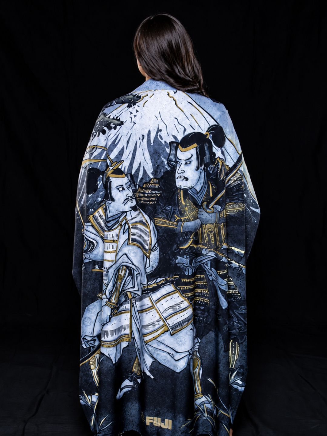 A person with long brown hair is draped in the Womens Heritage Keisho Lightweight BJJ Gi Black, which features illustrated samurai warriors in armor and mountains on a striking black backdrop.