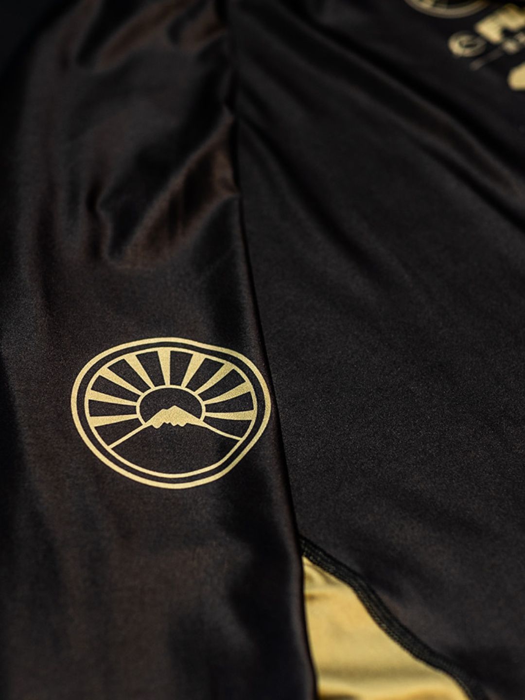A close-up of the Heritage Keisho Long Sleeve Rashguard Black, featuring a smooth, shiny black fabric with gold accents along the seam and a gold emblem depicting a stylized mountain and sun rays.