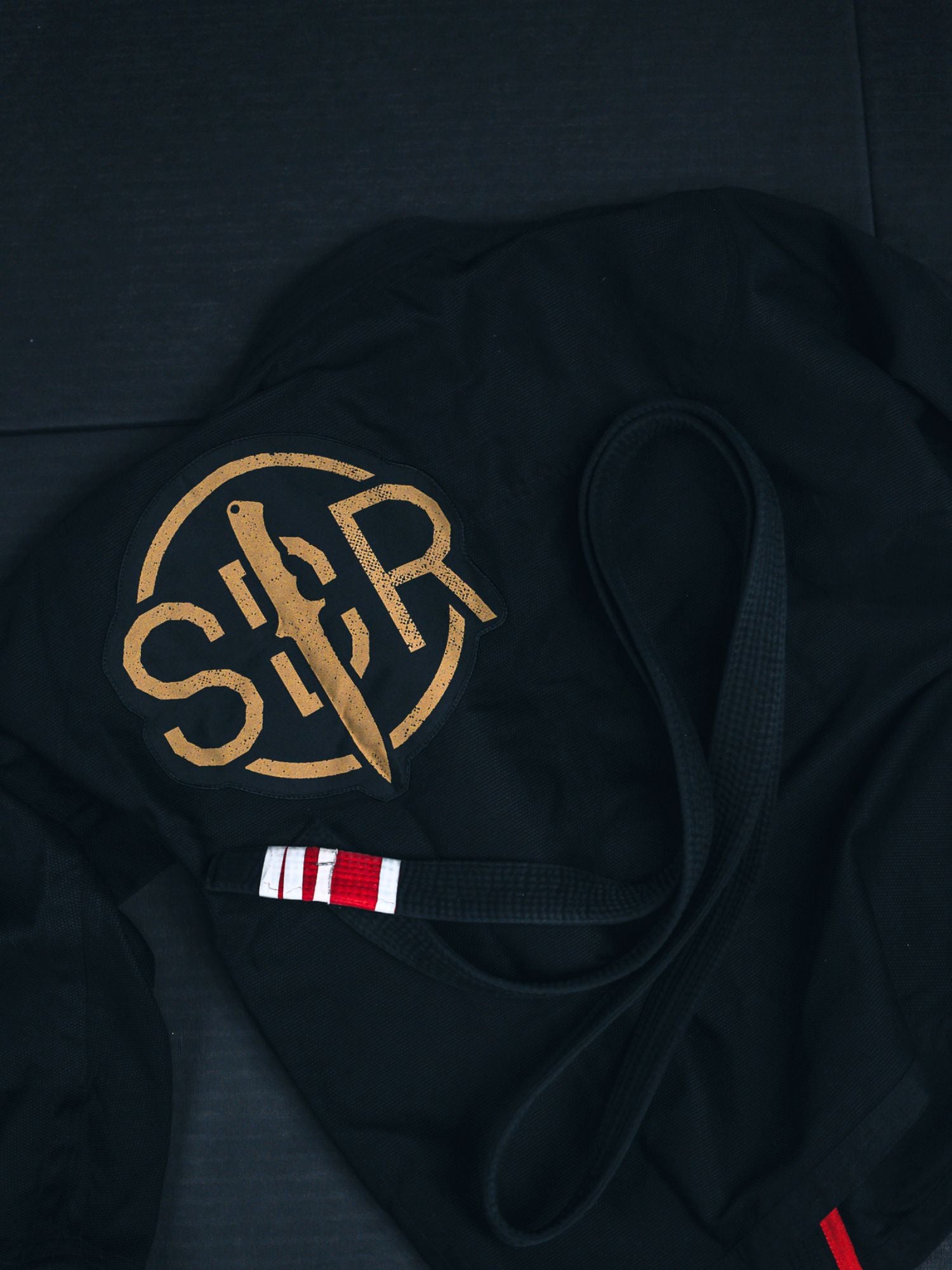 The Sheepdog Response Standard Suparaito BJJ Gi in black, featuring a gold SR logo and a black belt with red and white stripes, rests on a dark mat.