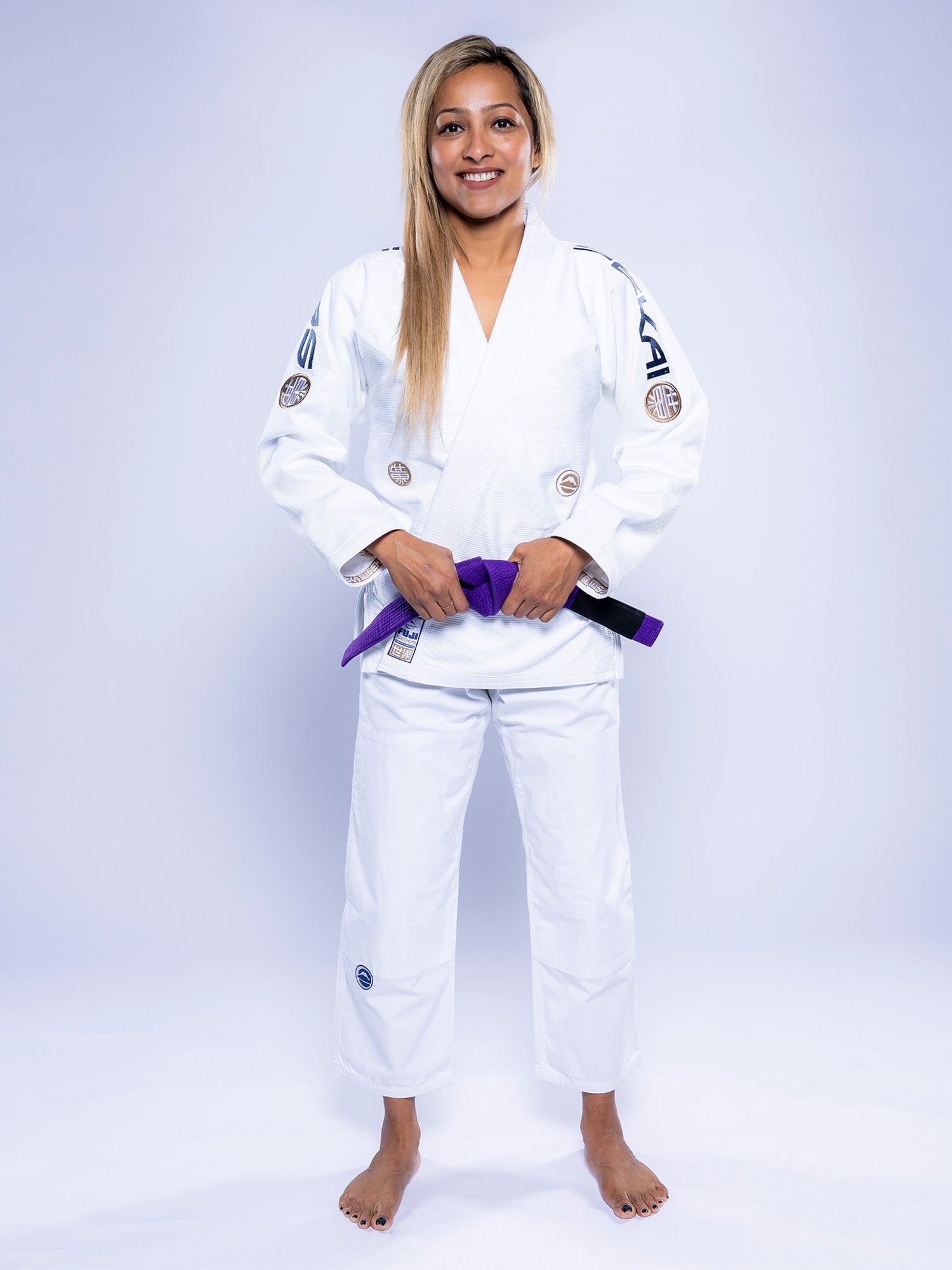 A woman wears the Womens SEKAI 3.0 BJJ Gi White Navy, standing barefoot and smiling as she holds a purple belt in front of her against a plain, light background.