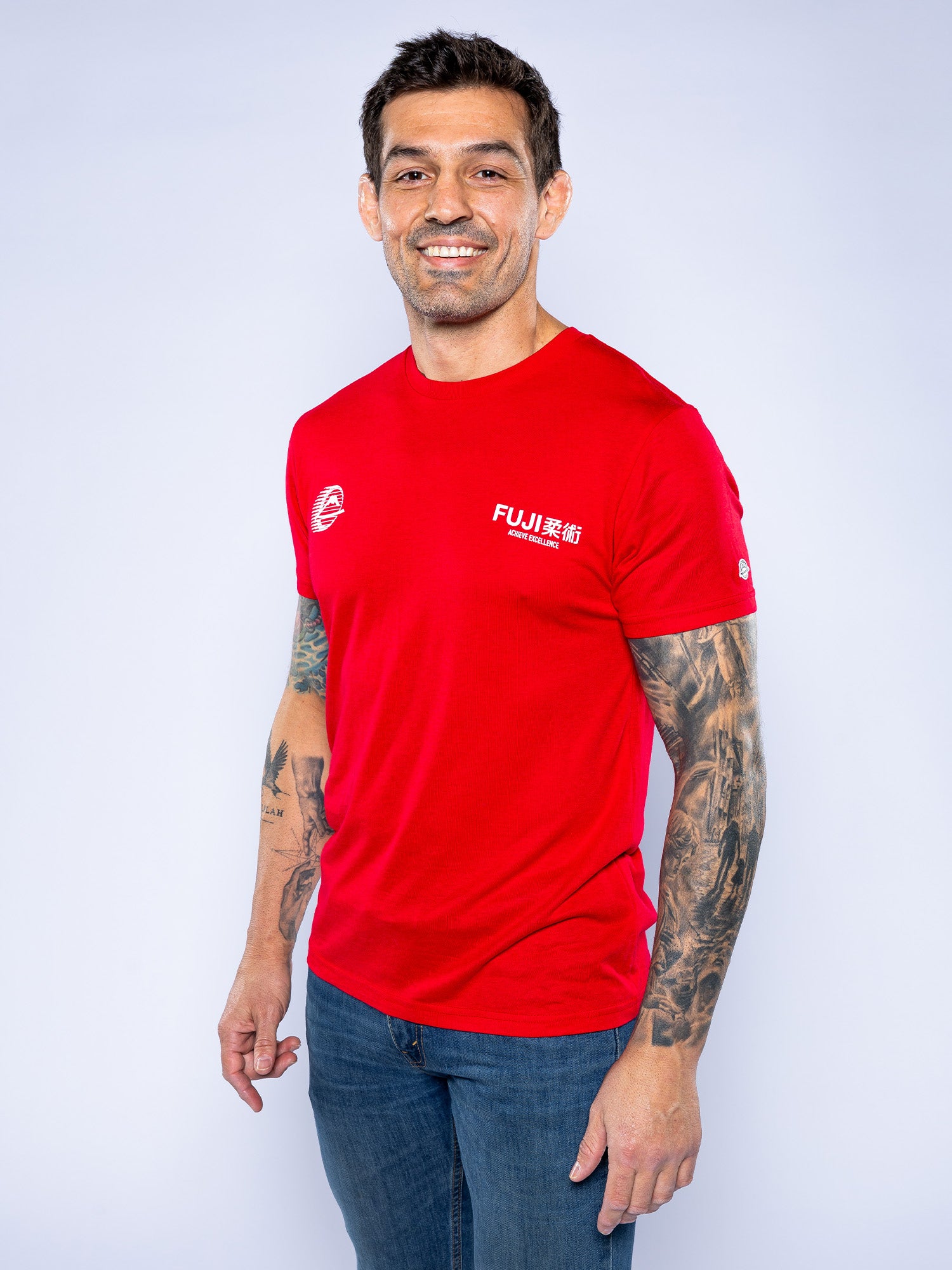 A man with tattoos on his right arm is smiling against a light background, wearing the bright red Iconic Jiu Jitsu Shirt and blue jeans.